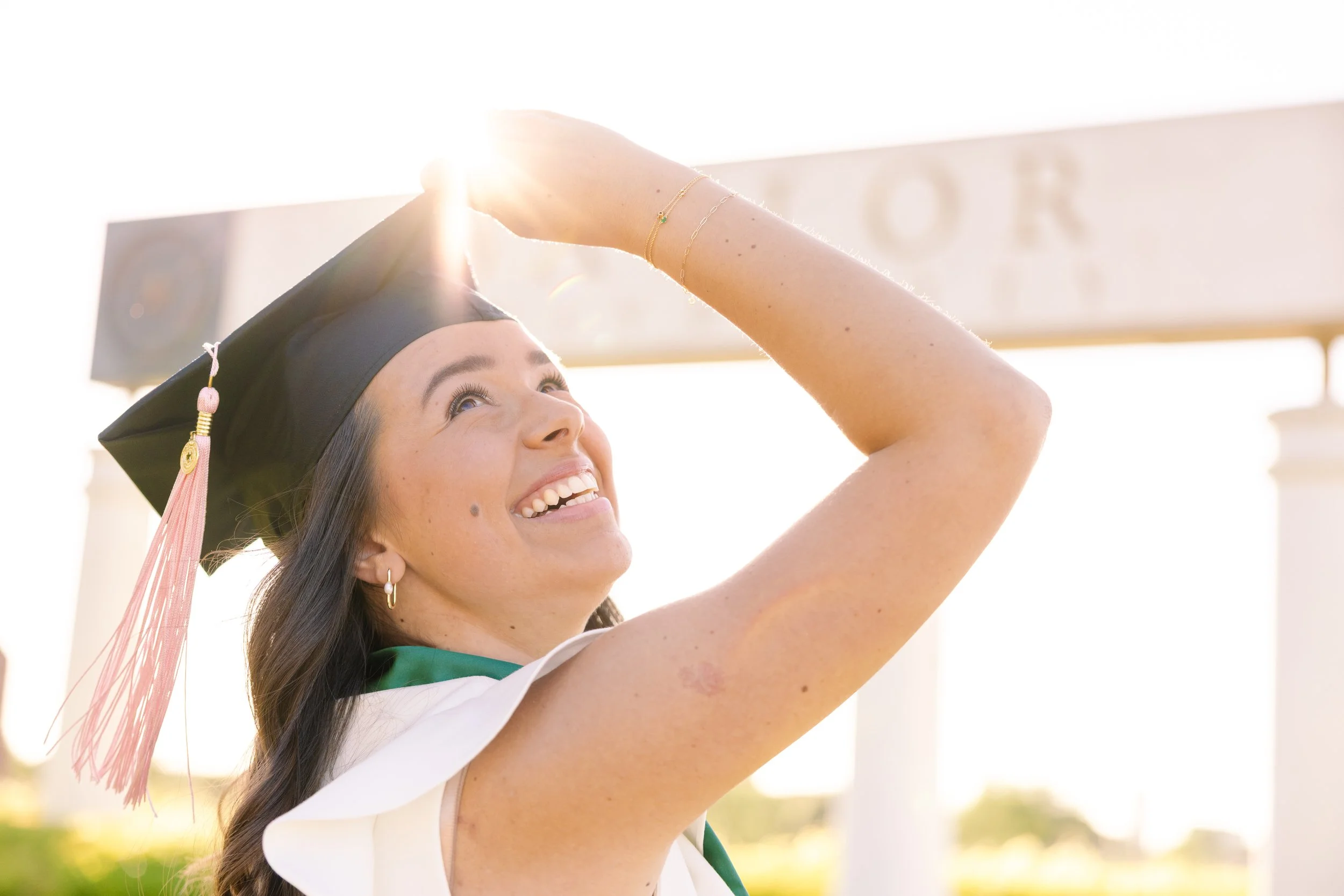 A woman in a graduation cap and gown smiling and adjusting her cap outdoors during the daytime.