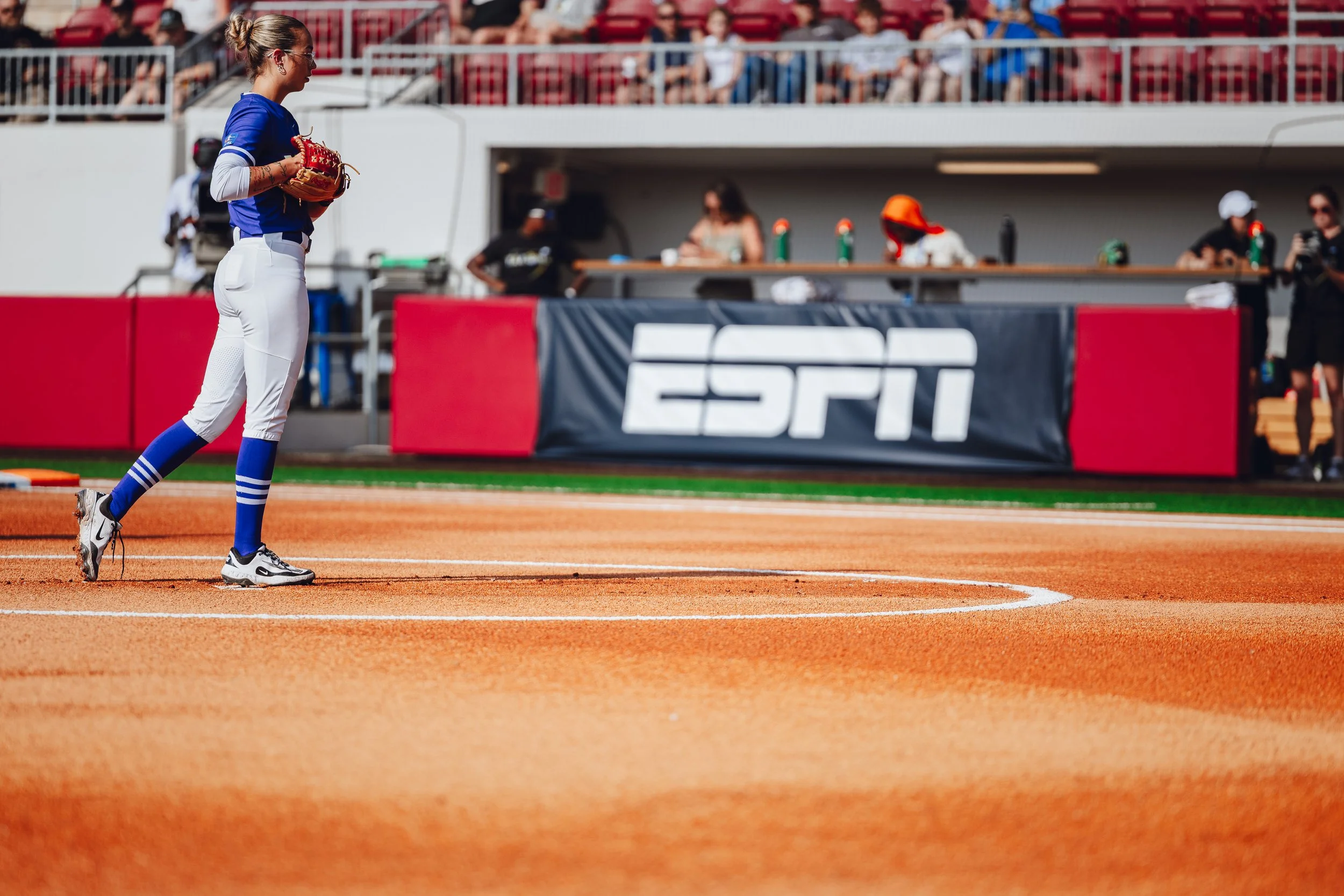 A female softball player standing on a dirt field with a glove, wearing a blue jersey, white pants, and blue and white socks at a stadium with ESPN signage in the background.