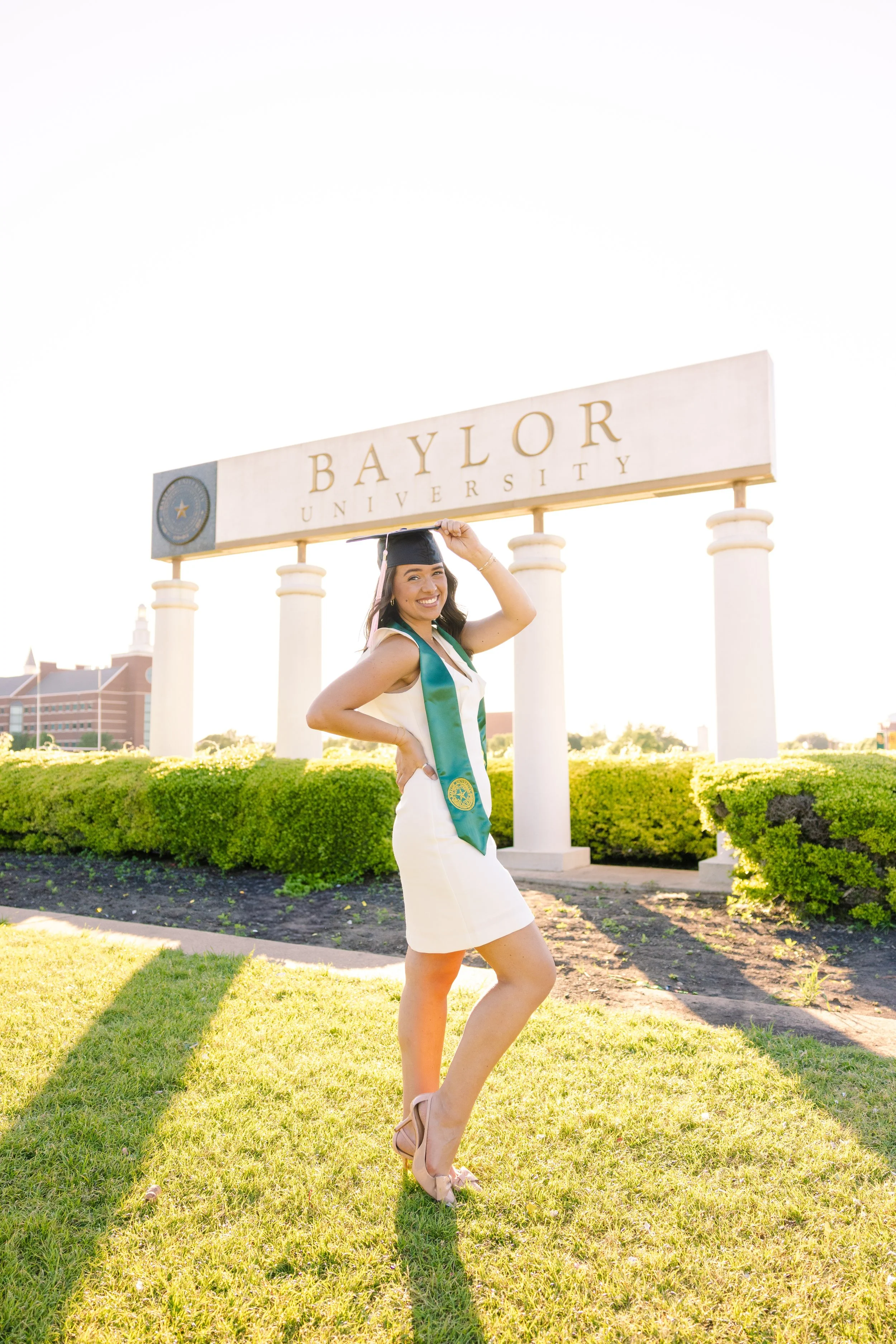 A woman in a graduation cap and gown with a Baylor University sash, standing on grass in front of a Baylor University sign during the daytime, smiling and adjusting her cap.