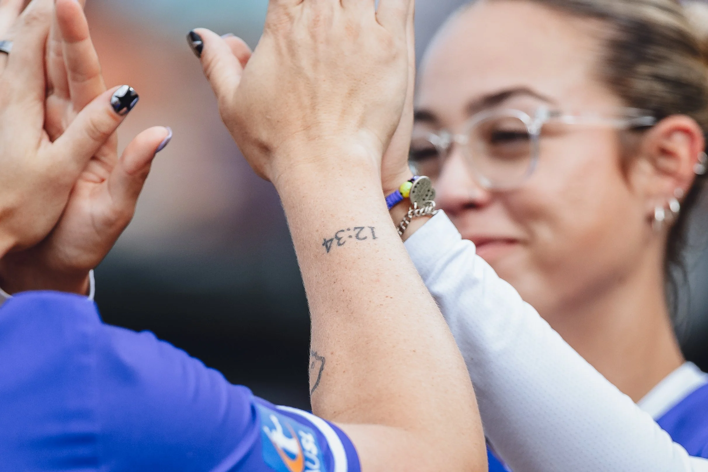 Two women high-fiving, one wears a blue jersey, and the other has glasses, earrings, and a tattoo on her arm.