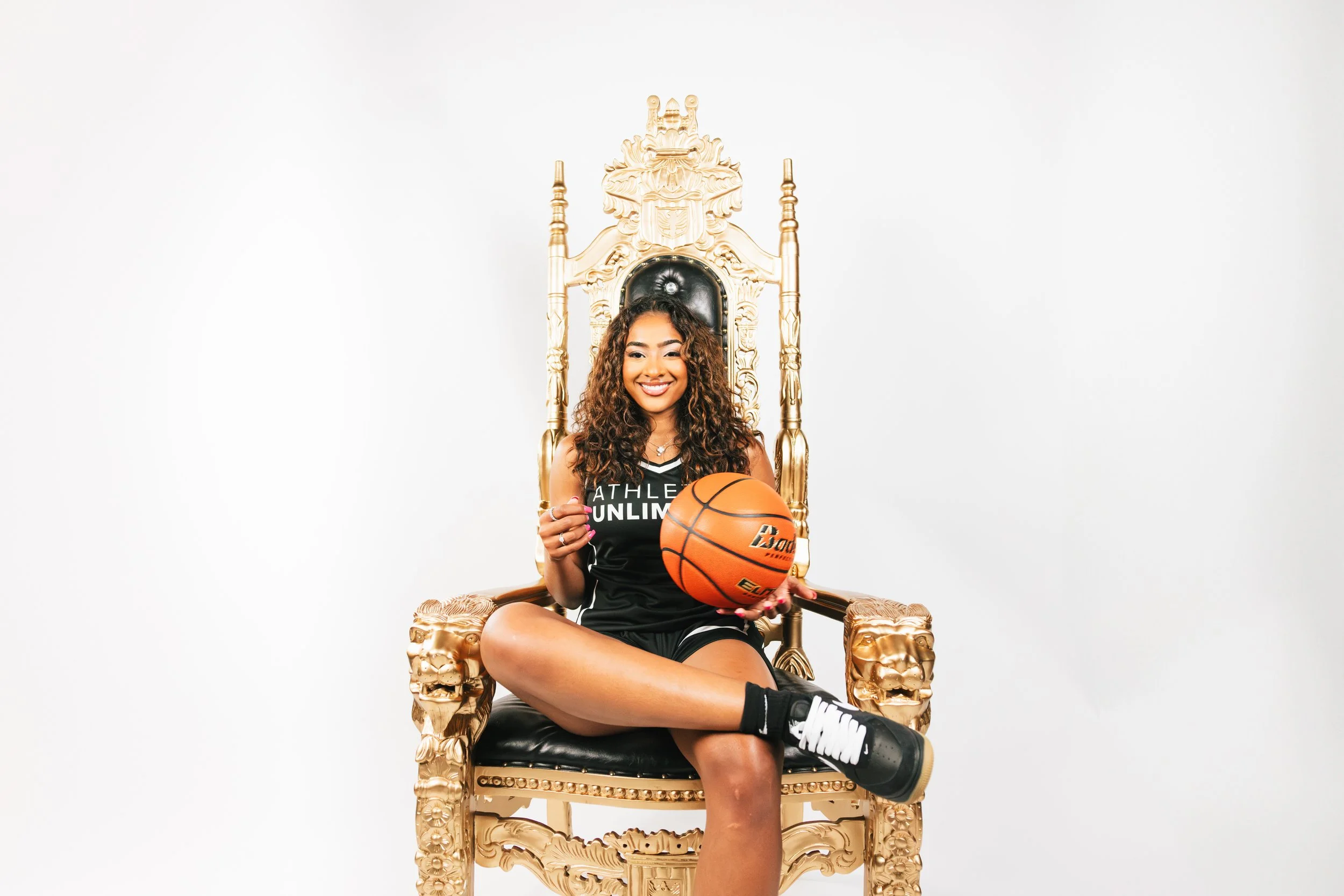 A young woman with curly hair sitting on a gold ornate throne-like chair, holding a basketball, smiling, wearing a black sports uniform with black sneakers, against a plain white background.