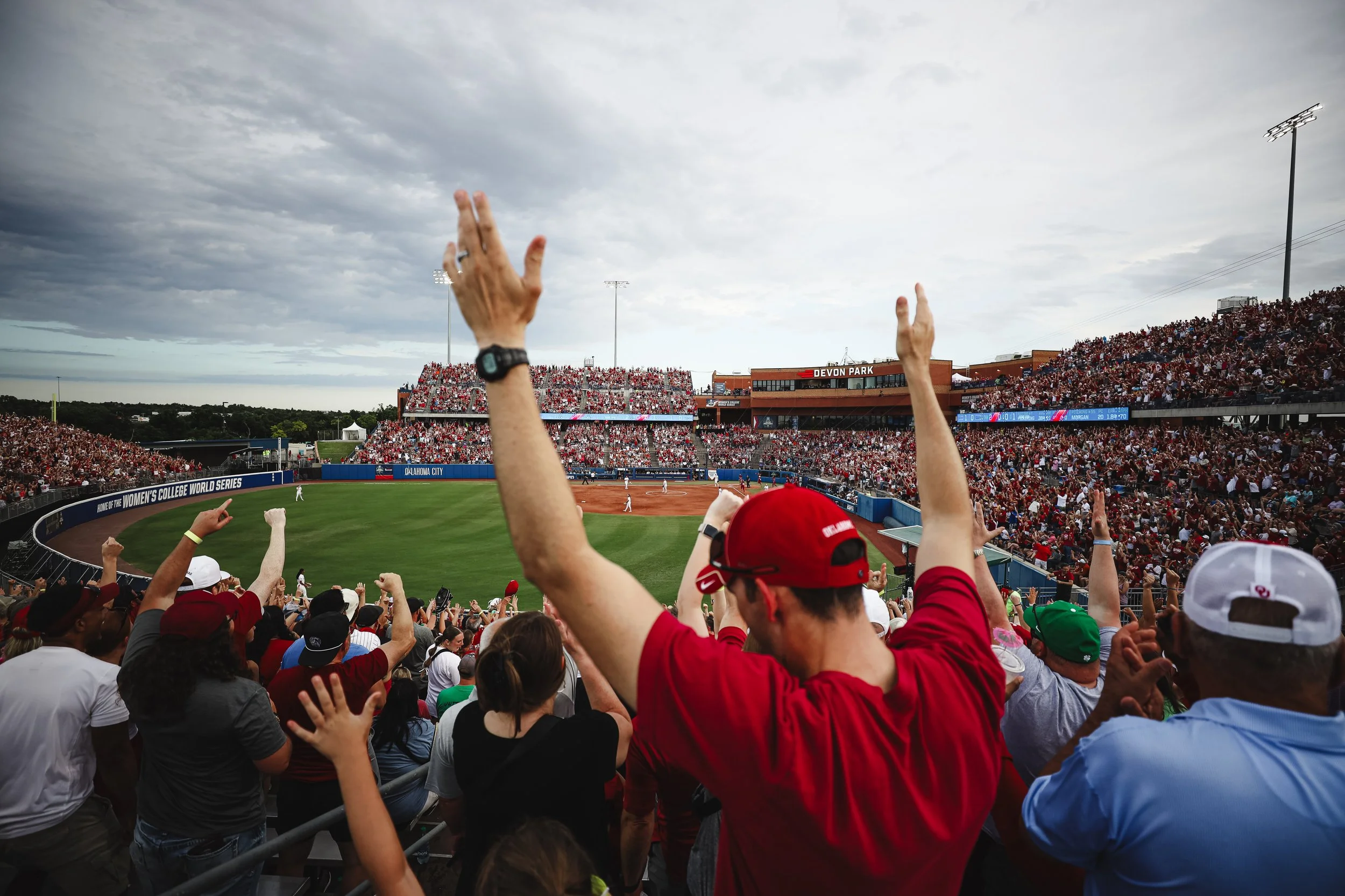 Crowd of Oklahoma fans cheering at a Women's College World Series softball game in a stadium with a view of the field and cloudy sky above.