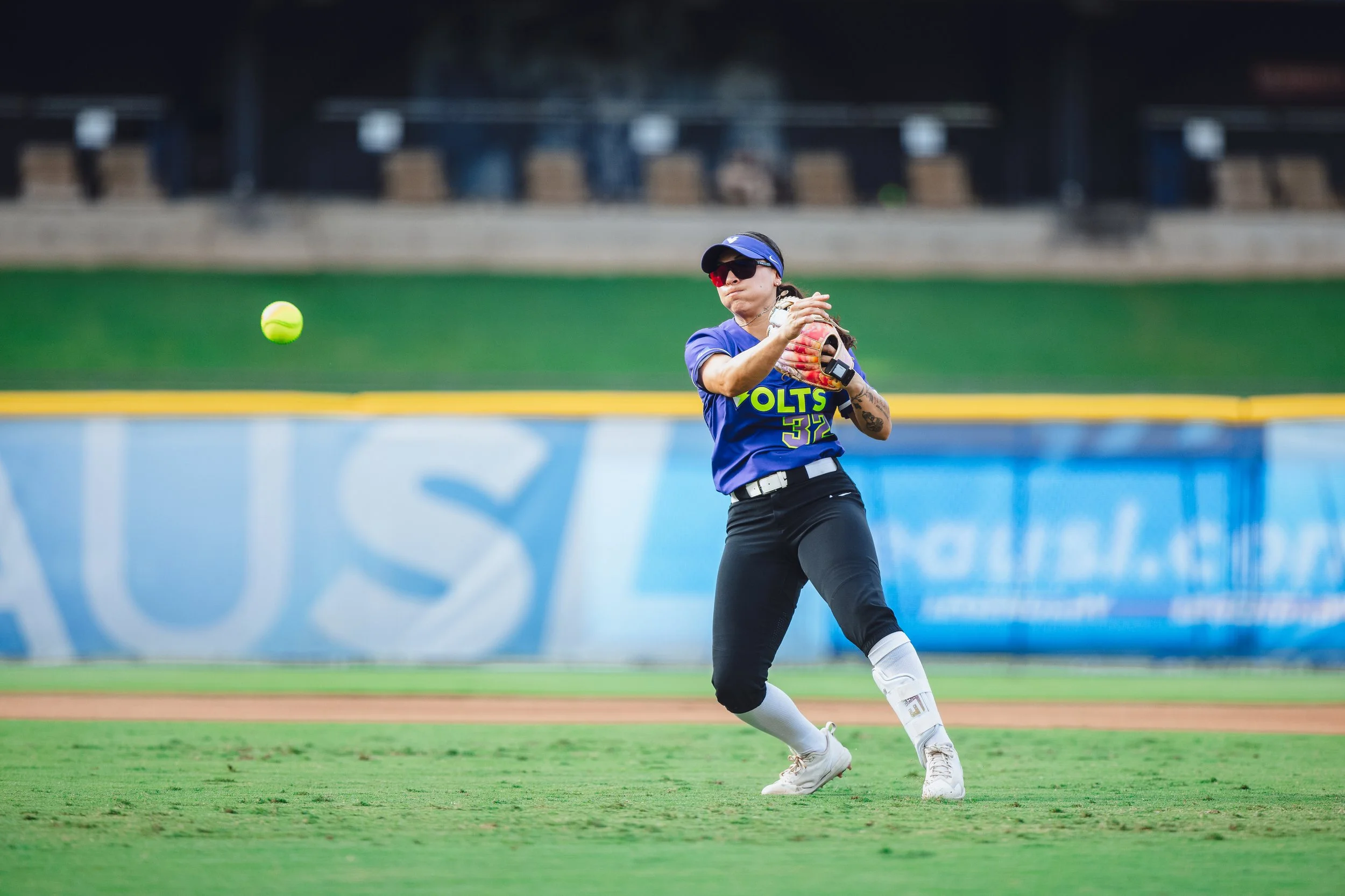 A female softball player in a blue jersey with yellow lettering, wearing black pants, white socks, and white cleats, is preparing to catch a bright green softball on the field.