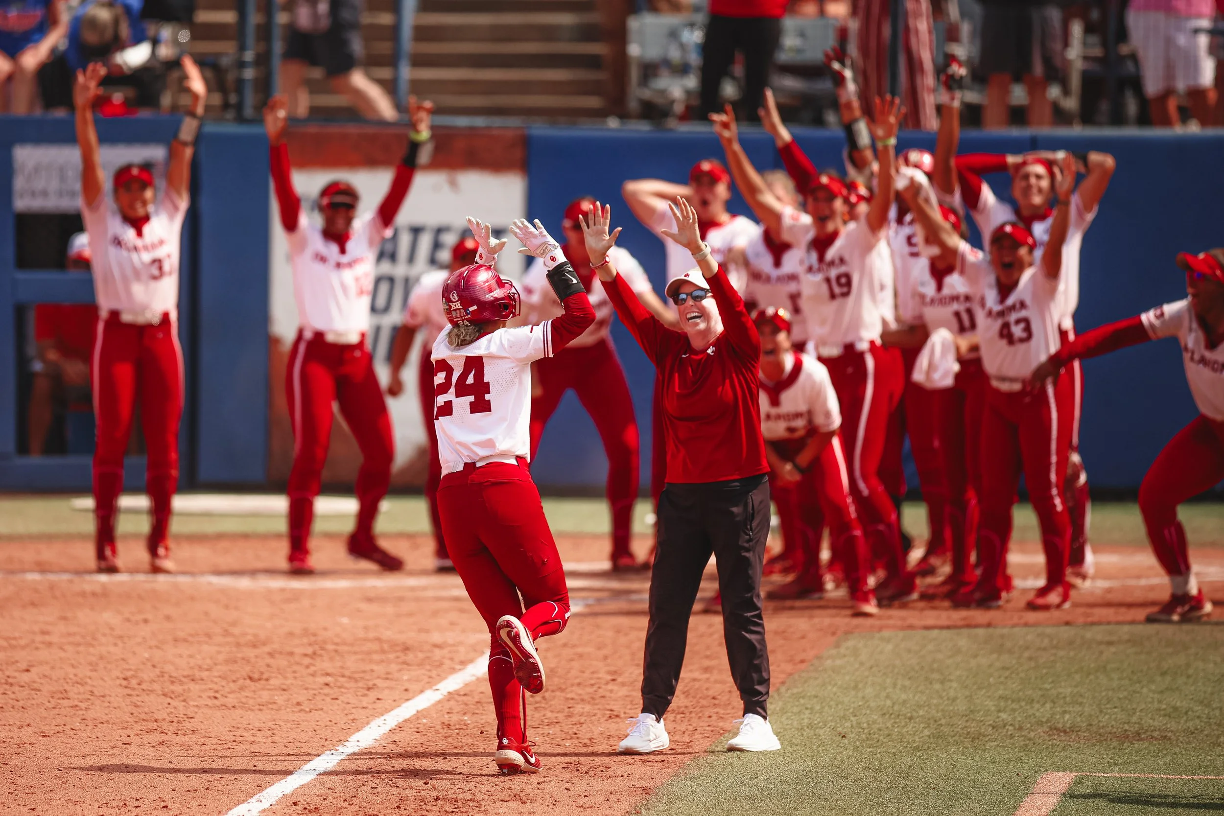 A female softball player high-fives a woman, possibly her coach, on the field near home plate during a game. In the background, a group of players and staff cheer and celebrate.