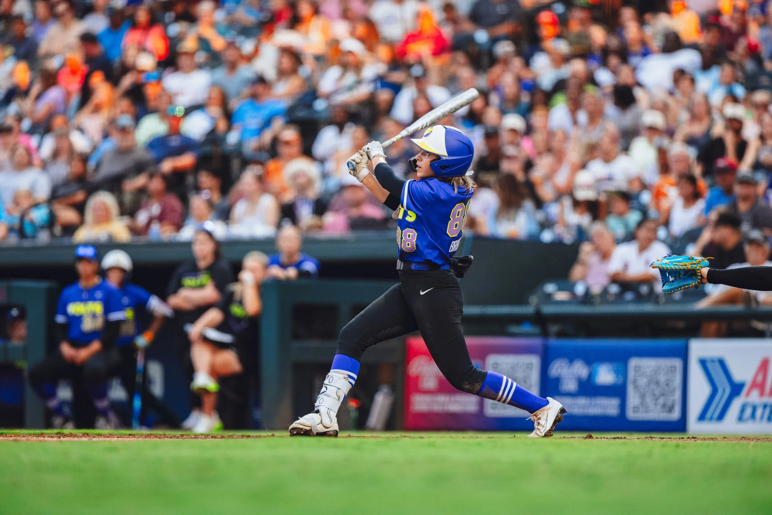 Female softball player in a blue jersey swinging a bat during a game, with an audience in the background.
