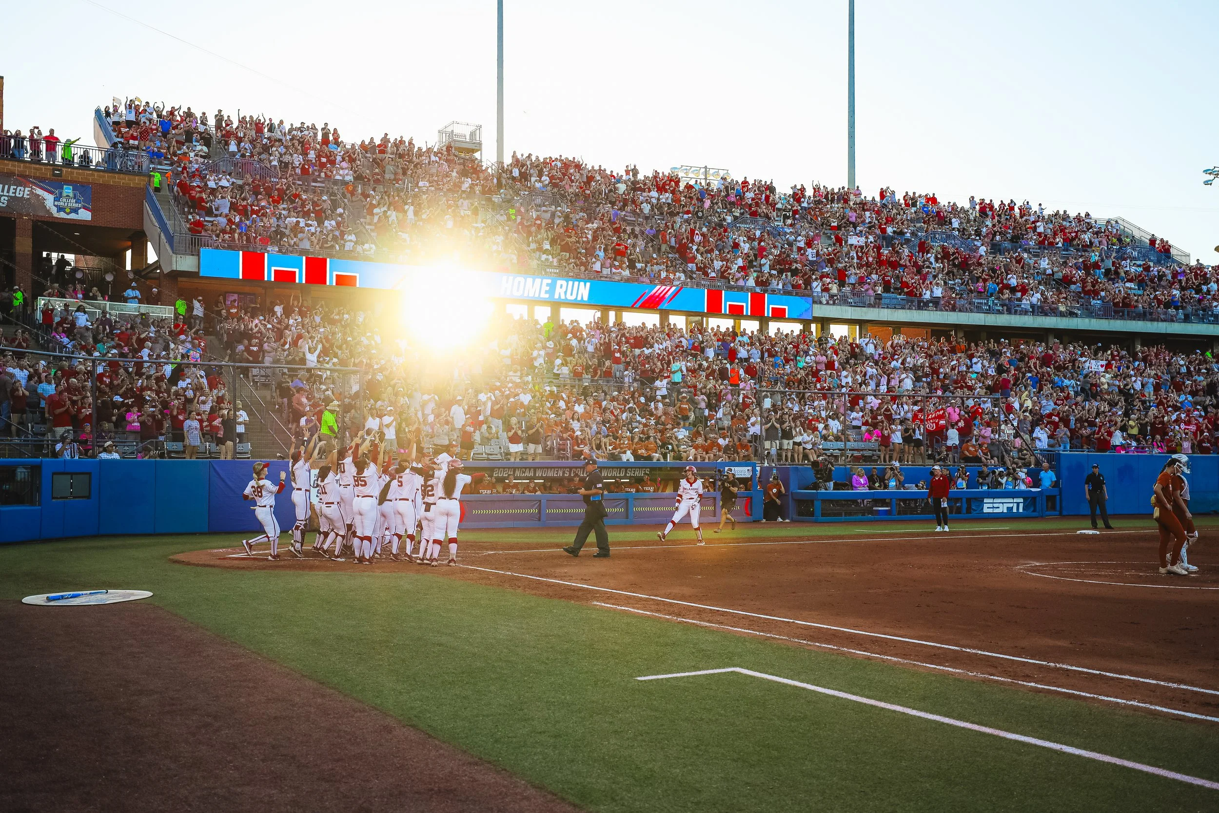 A baseball team celebrating a home run at a stadium with a large crowd watching, and bright sunlight in the background.