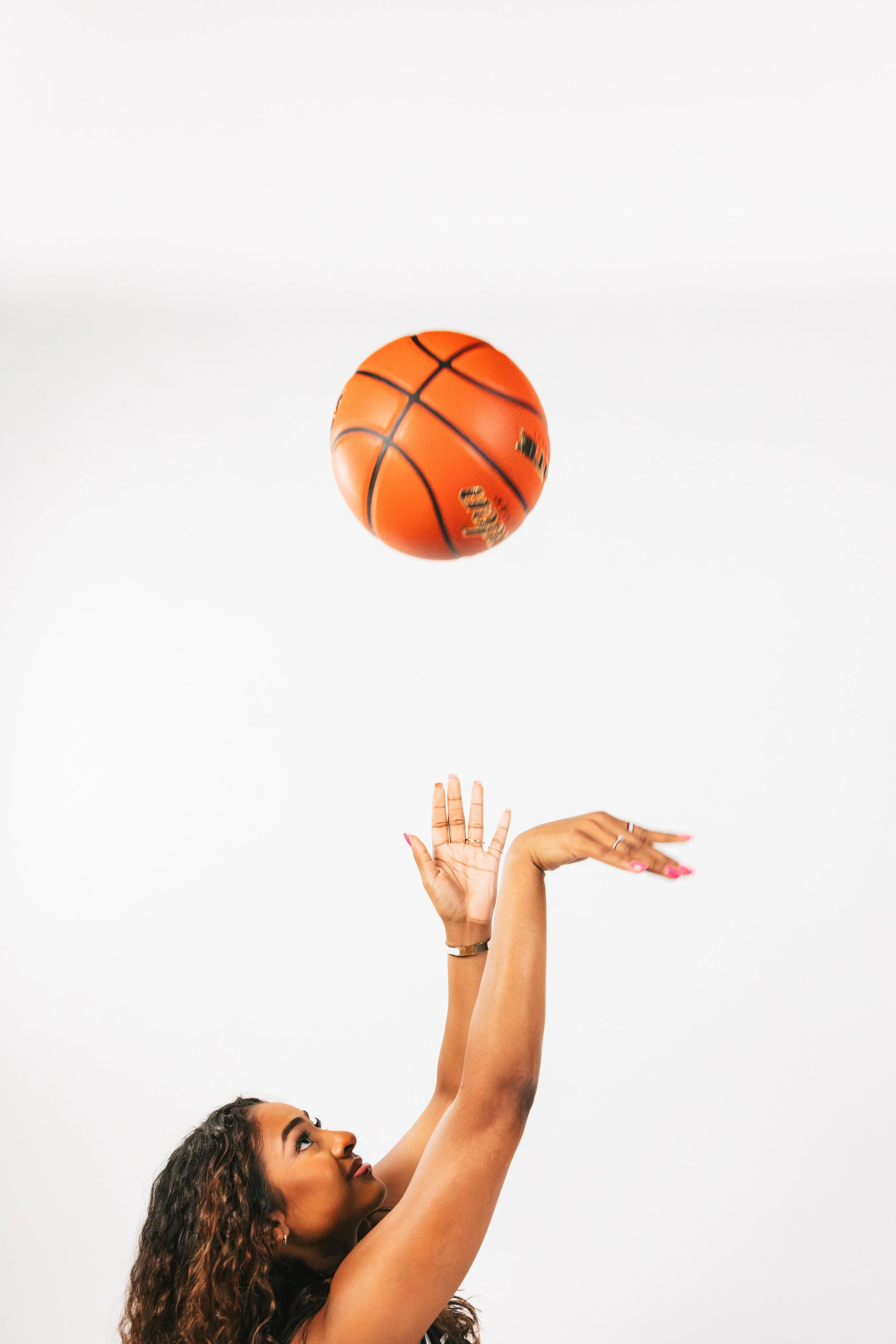 A woman with curly hair reaching up to catch or hit an orange basketball against a white background.