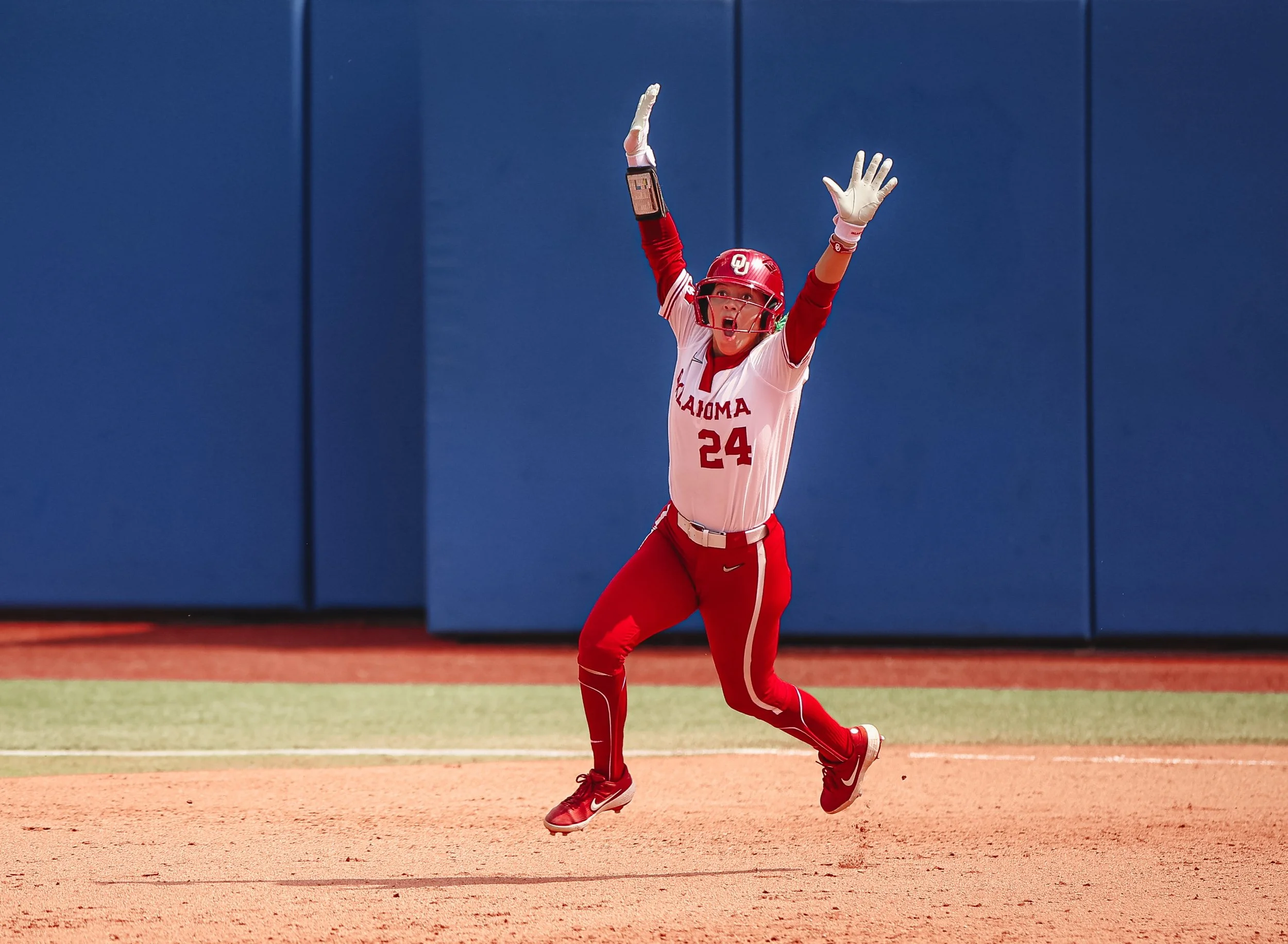 A female softball player in a white and red uniform with the word "Oklahoma" and the number "24" is celebrating on the field with arms raised and mouth open.