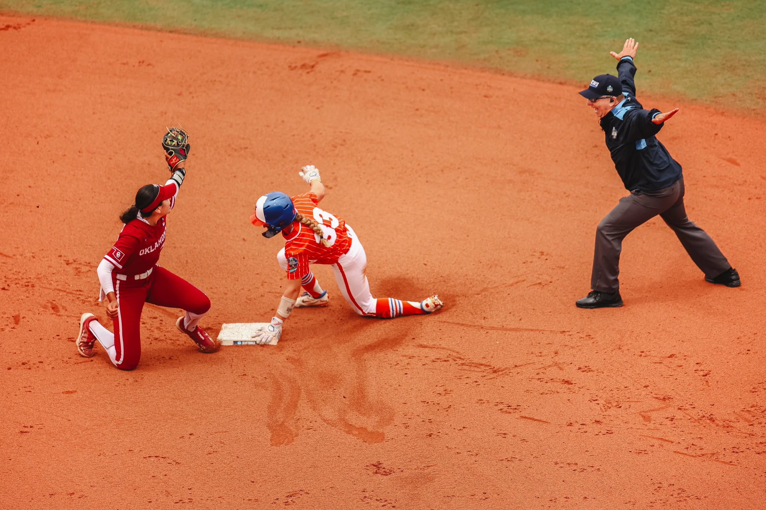 Young baseball player in a red and orange uniform slides into a base as an umpire calls the play, while a teammate in a red uniform knees nearby on the reddish dirt field.