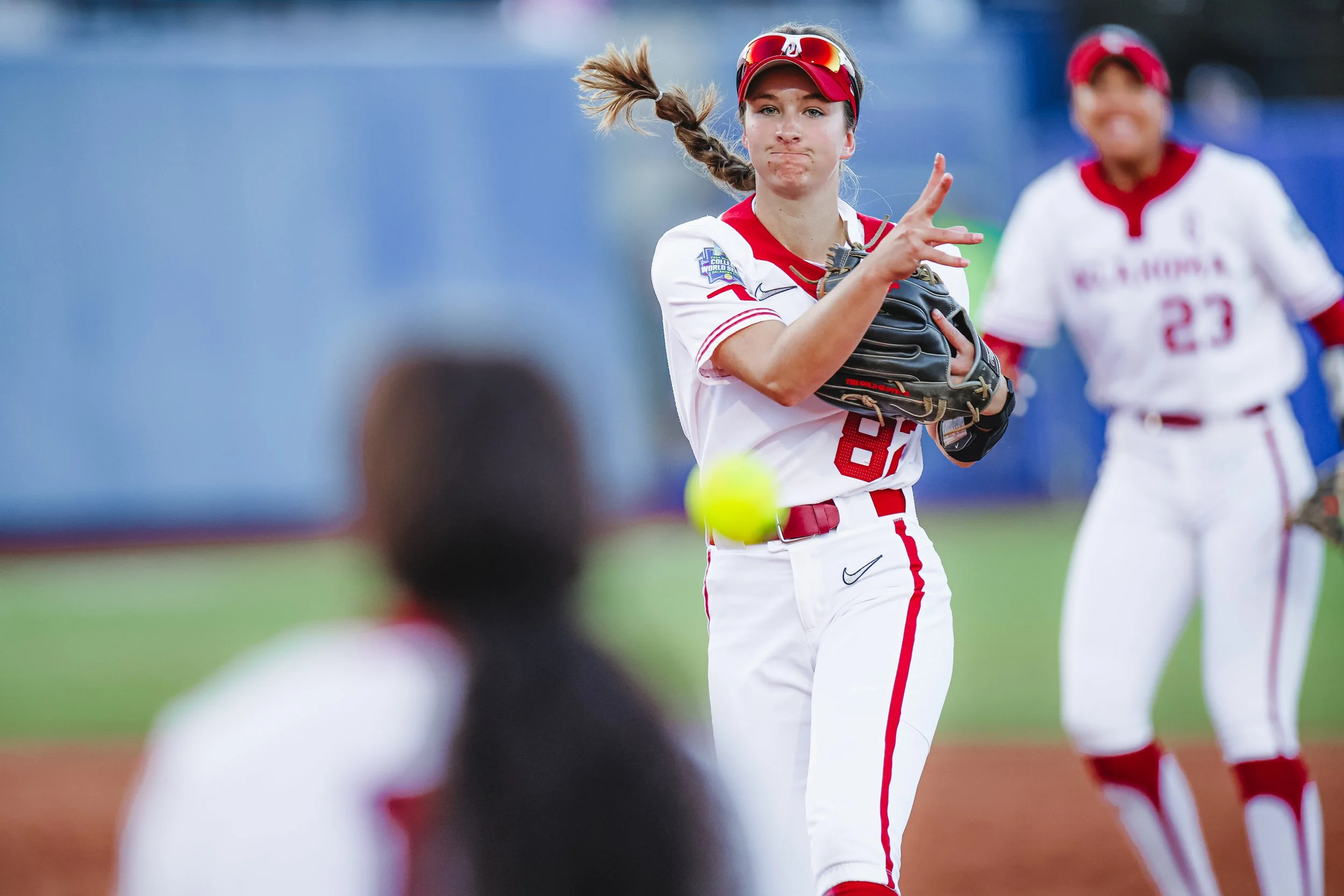 A female softball player in a white and red uniform catches a yellow softball with her glove on a field, with a teammate in similar uniform in the background.