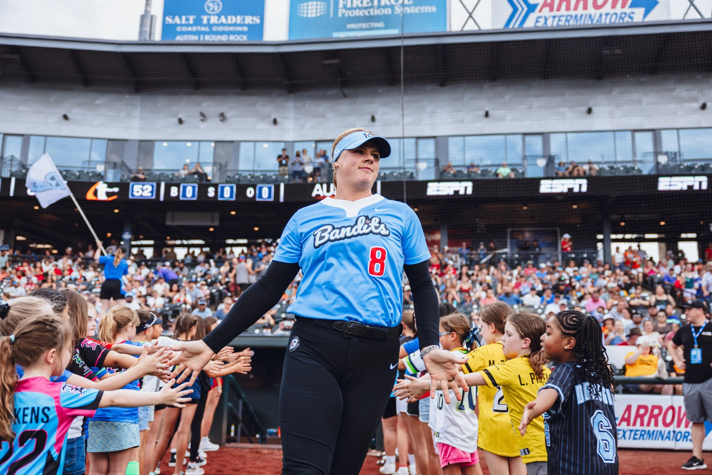 A woman in a blue sports jersey with the number 8 and the word 'Bandits' on it, standing on a baseball field high-fiving young girls as part of a ceremony, with a large crowd in the stands and a scoreboard in the background.