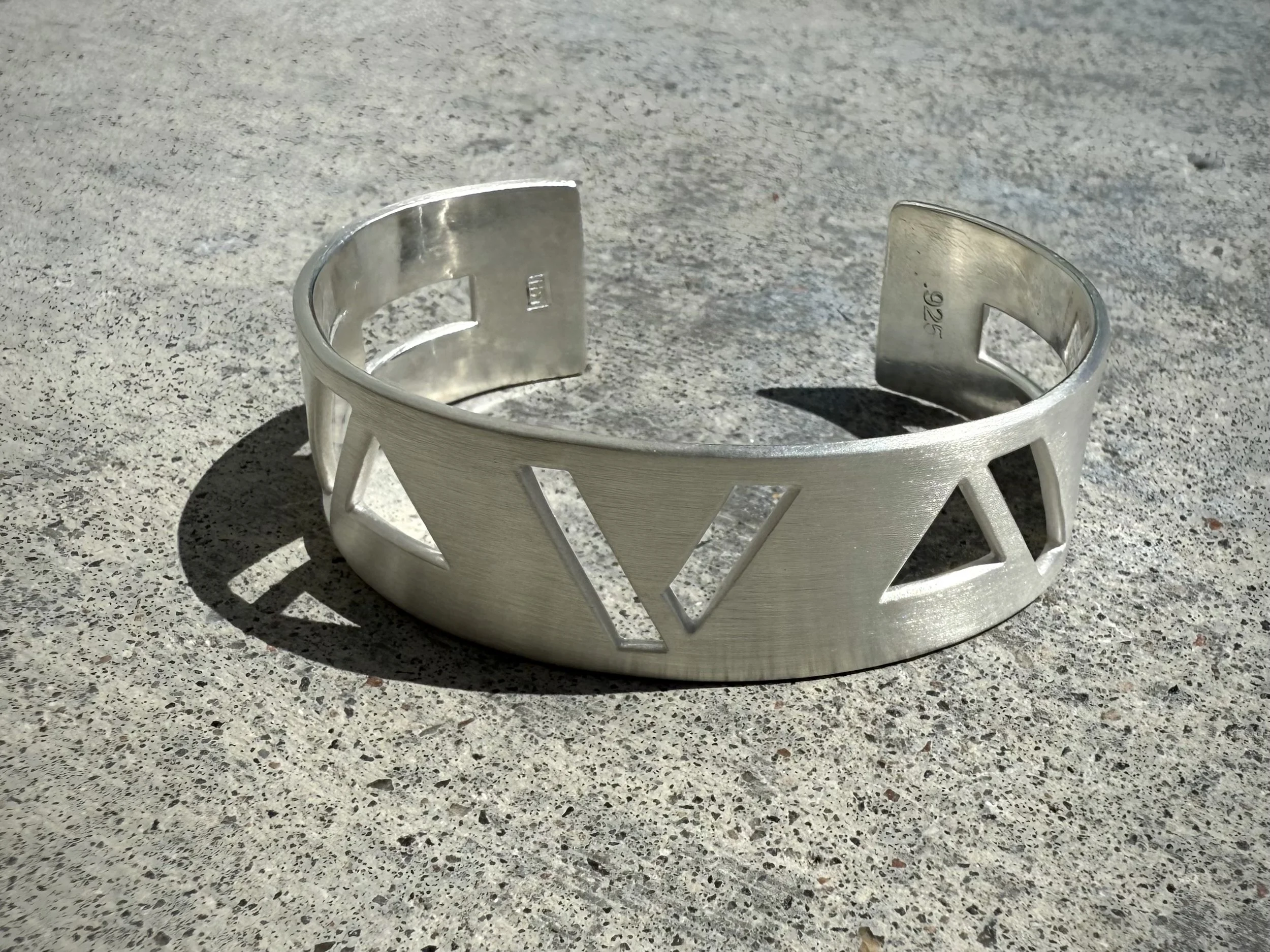 A silver metal cuff bracelet with cut-out triangle patterns resting on a gray textured surface.