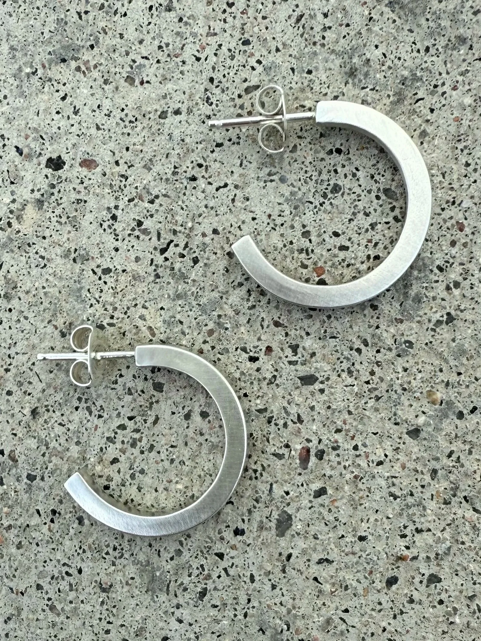 Silver hoop earrings with post backs laying on a speckled gray surface.