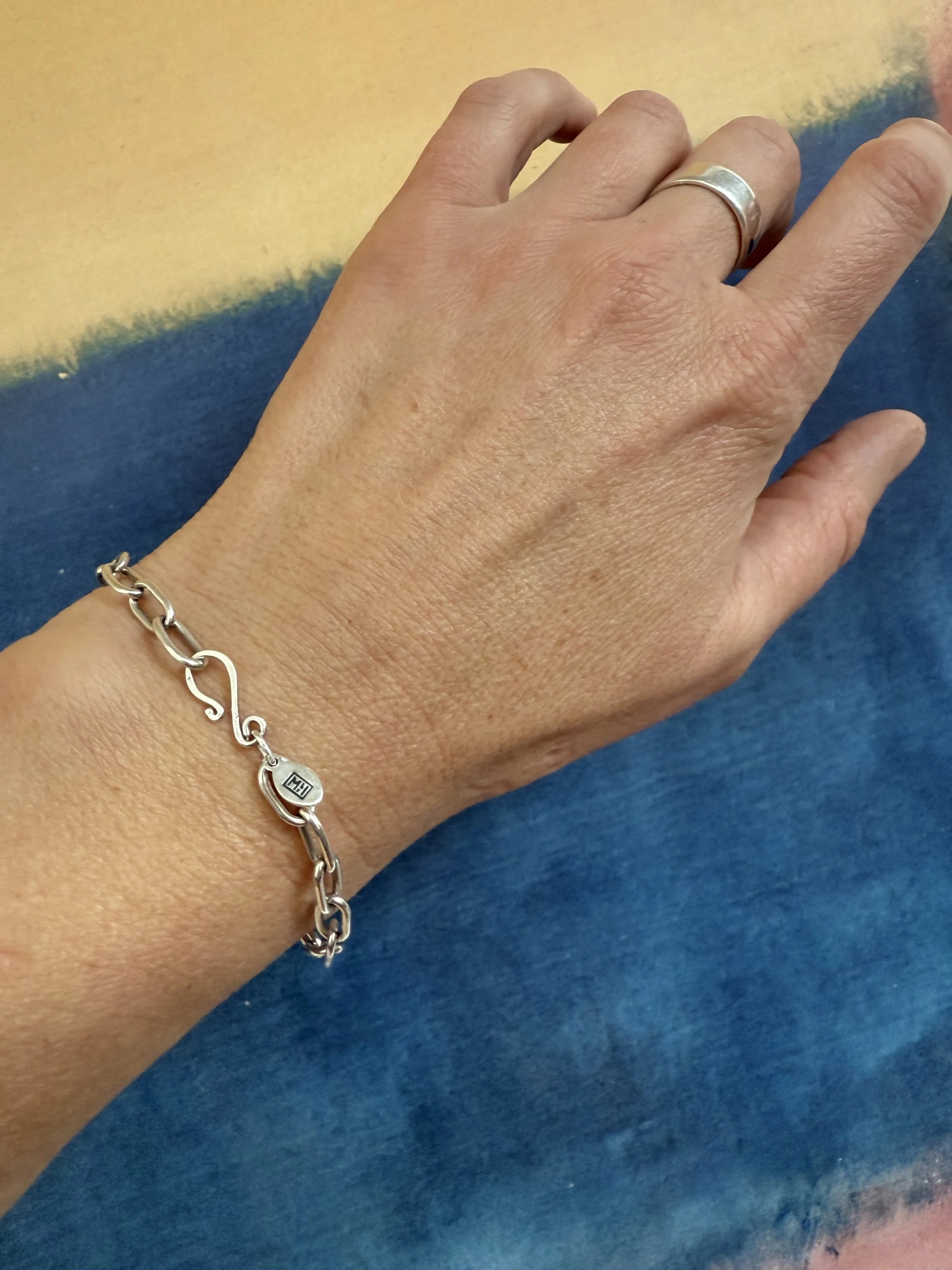 Close-up of a person's left hand wearing a silver ring on the ring finger and a silver bracelet with a small tag stamped with the MLH Designs logo around the wrist, placed against a colorful background.