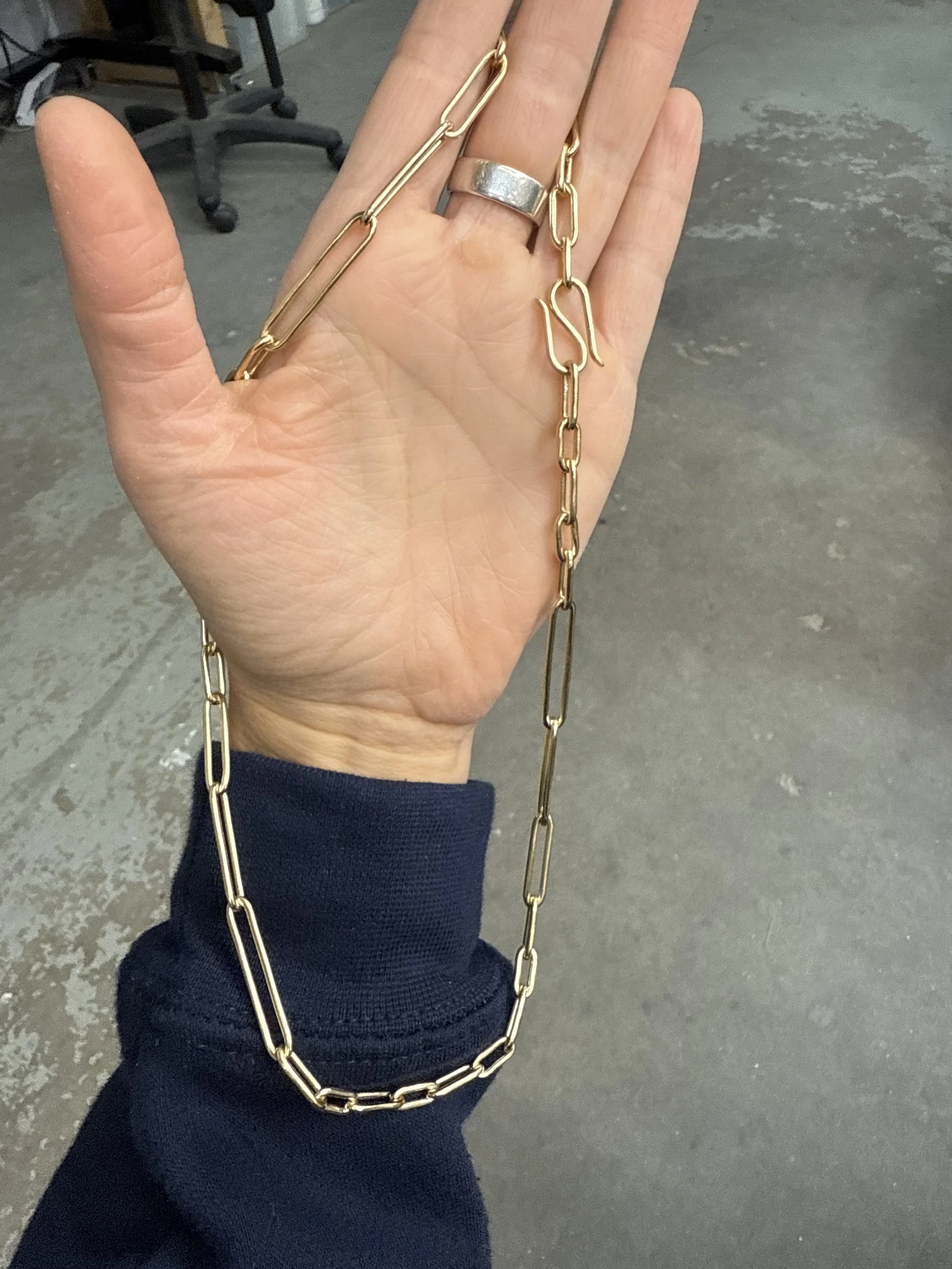 Hand holding a brass chain necklace and a silver ring, with a grey concrete floor in the background.