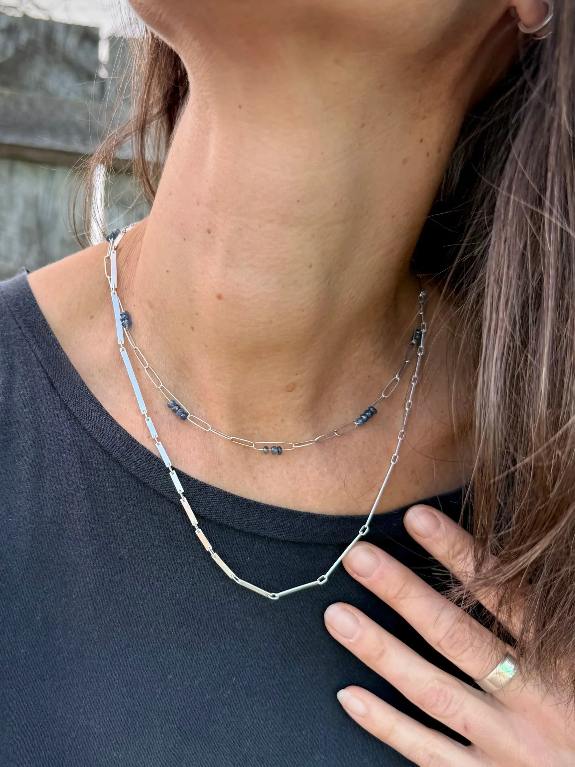 Close-up of a woman's neck and chest area, wearing two layered silver chain necklaces, one with small dark blue (3mm Kyanite faceted round) beads, and a silver ring on her finger, with a black shirt and outdoor background.