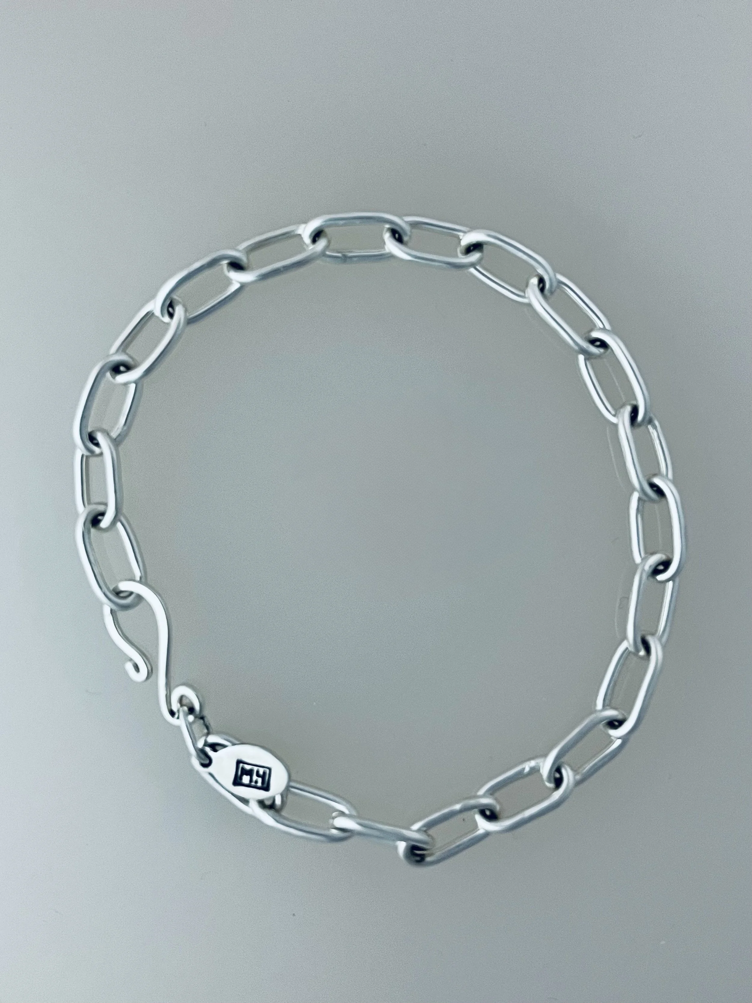 Silver chain bracelet with a small tag stamped with the MLH Designs logo and a handmade clasp, arranged in an open oval shape on a plain background.