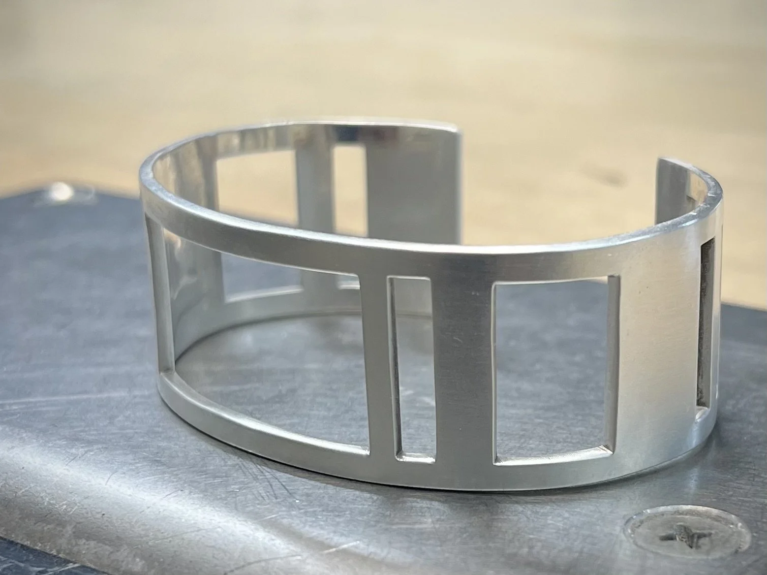 A silver metal cuff bracelet with rectangular cutouts is placed on a metallic surface.