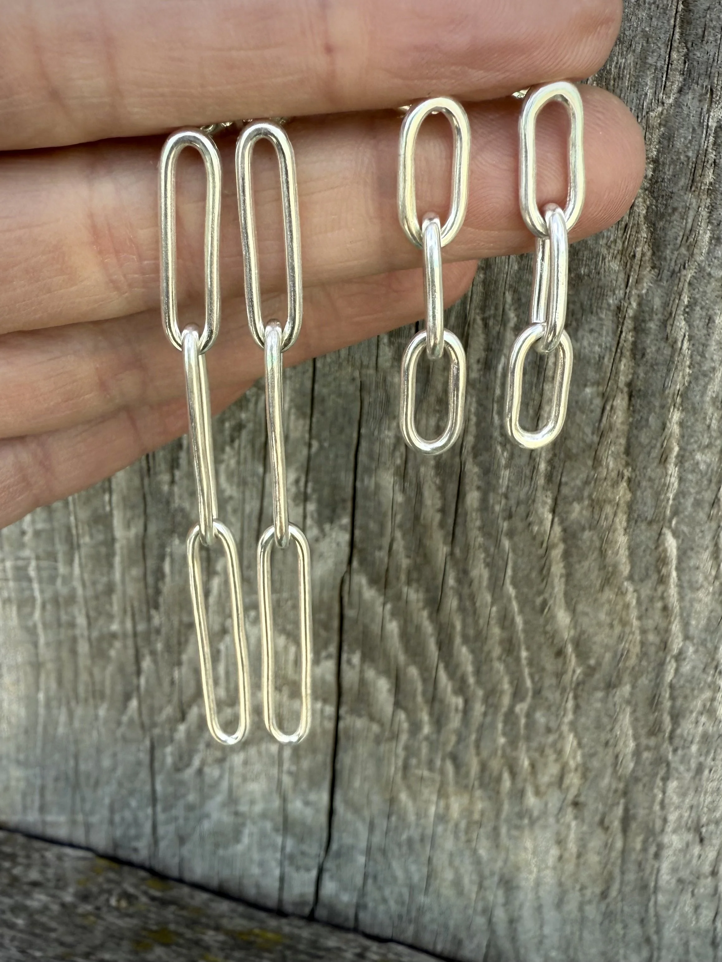 Two pairs of silver chain link earrings hanging from fingers against a wooden background.