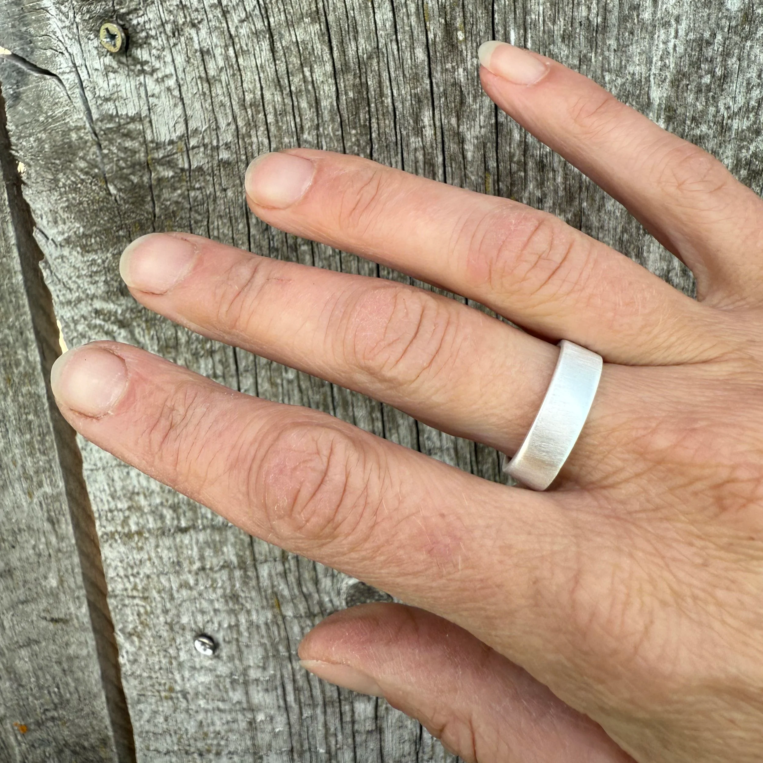 A hand with a wide band sterling silver ring on the middle finger resting on a weathered wooden surface.