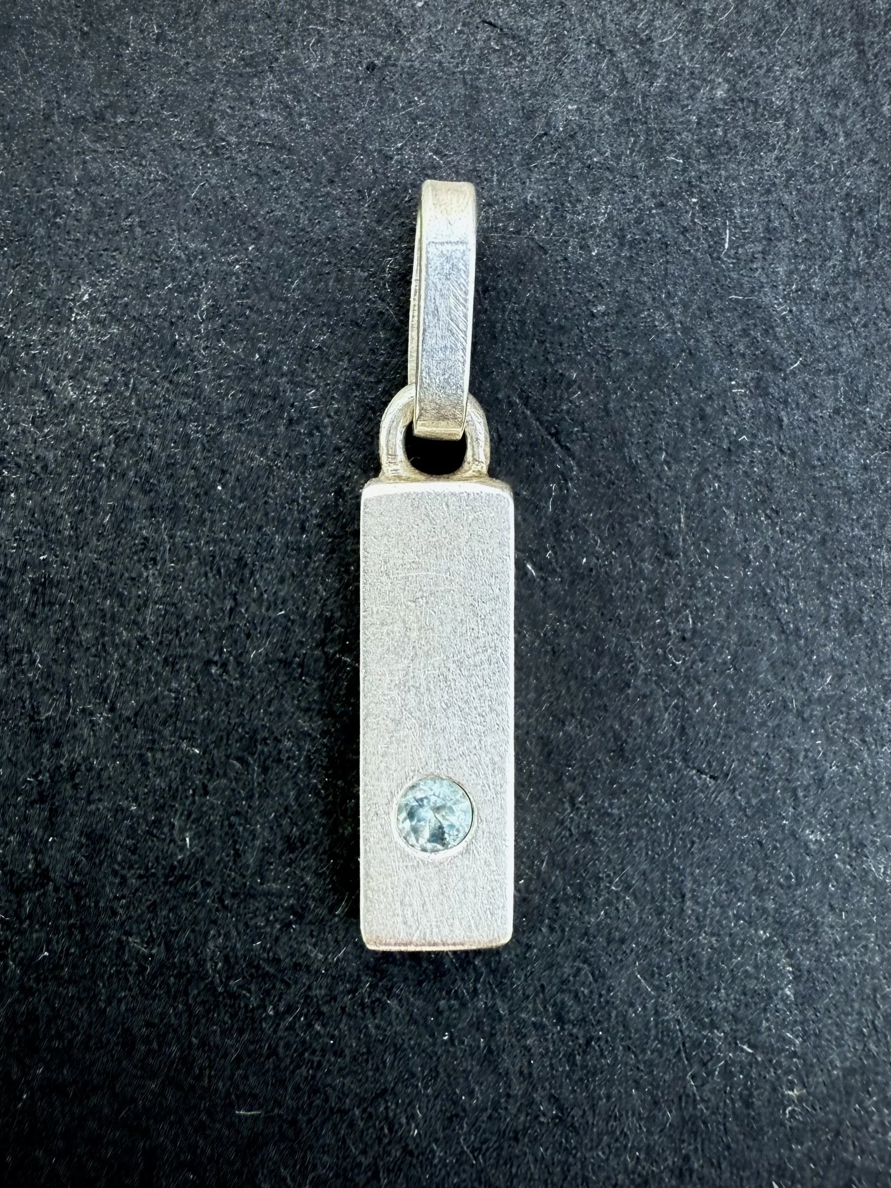 A silver pendant with a small gemstone, attached to a metal loop, placed on a black textured surface.