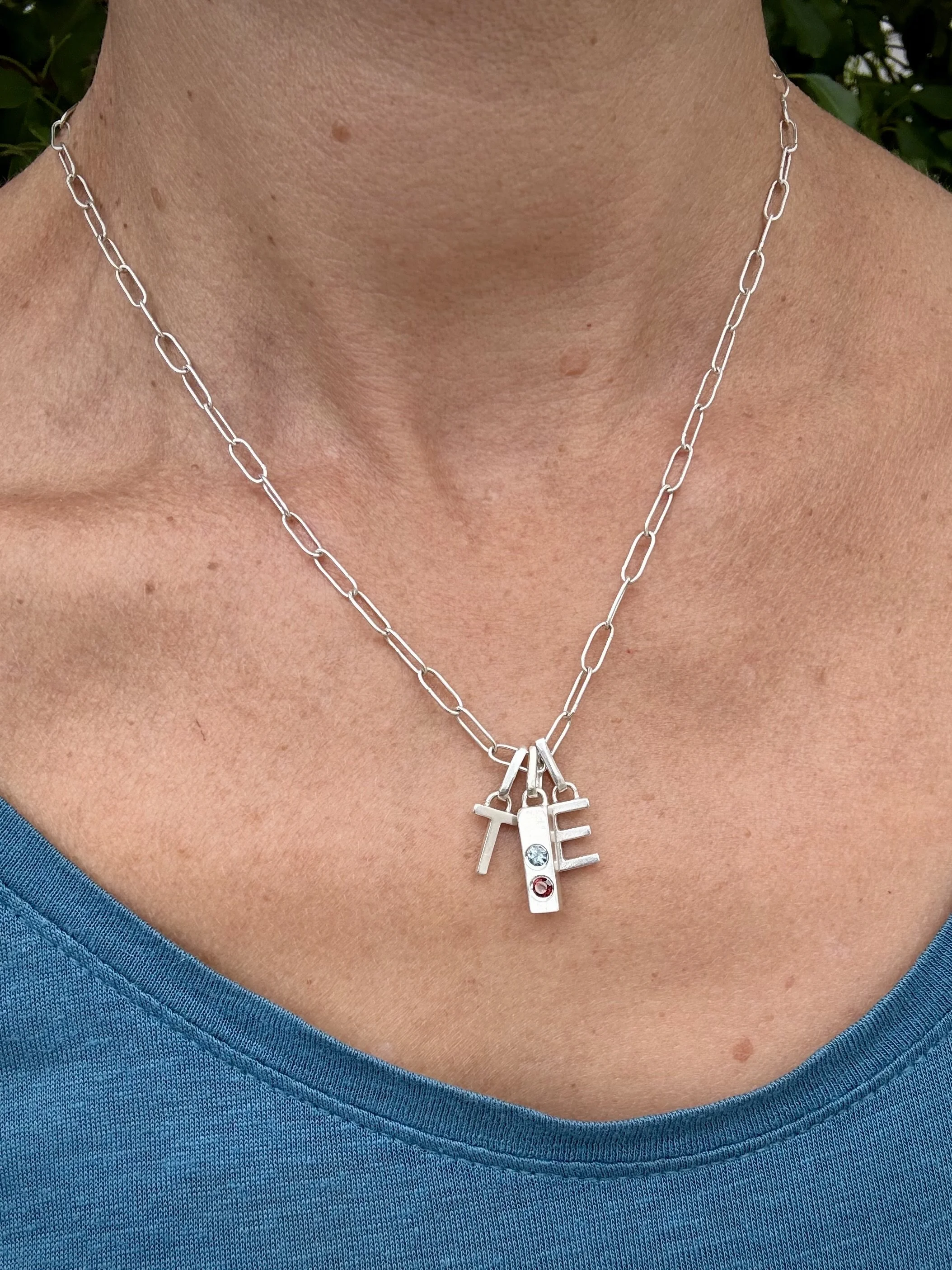 Silver necklace with two Capital Letter pendants ('T' and 'E'), and a silver bar pendant with a flush-set 2.5mm Montana Sapphire, worn on a person's neck over a blue top.