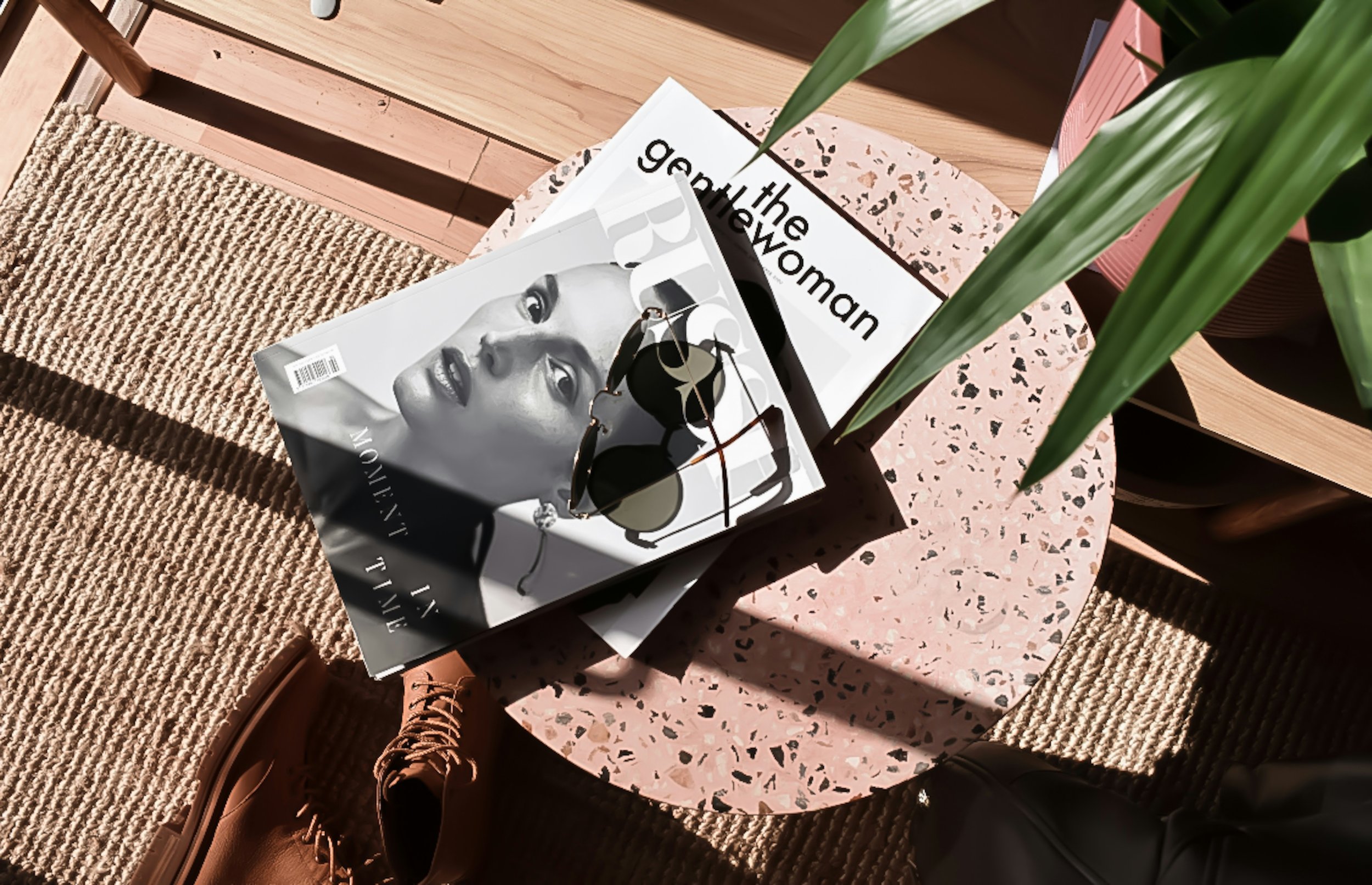 Magazines on a terrazzo table, sunglasses, brown boots, and a green plant.