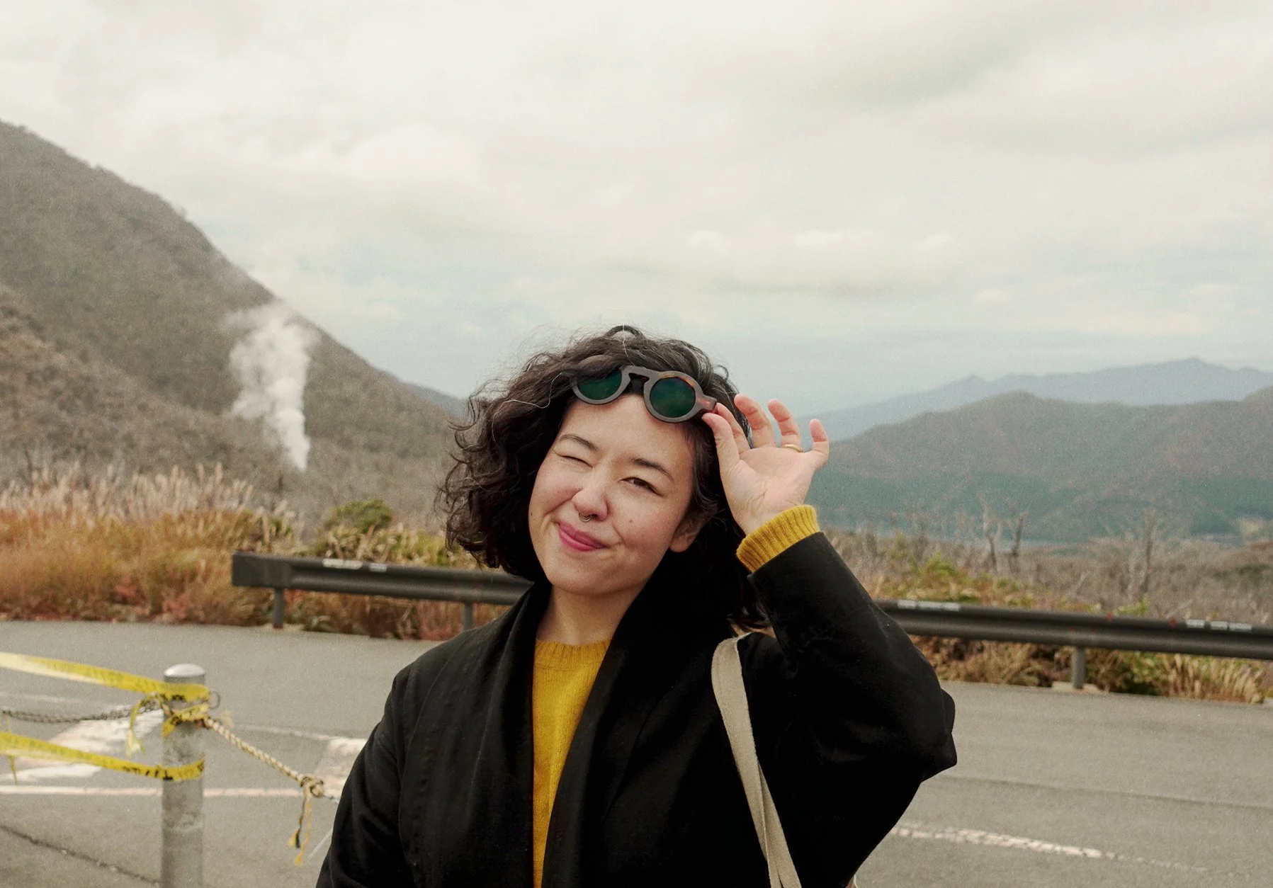 A young woman with curly dark hair winks and smiles at the camera while holding sunglasses on her head. She is wearing a yellow sweater and a black jacket with a beige bag strap. The background features mountainous terrain, cloudy sky, and some steam or smoke rising from the mountains. There is a metal guardrail and a road with yellow caution tape in the foreground.