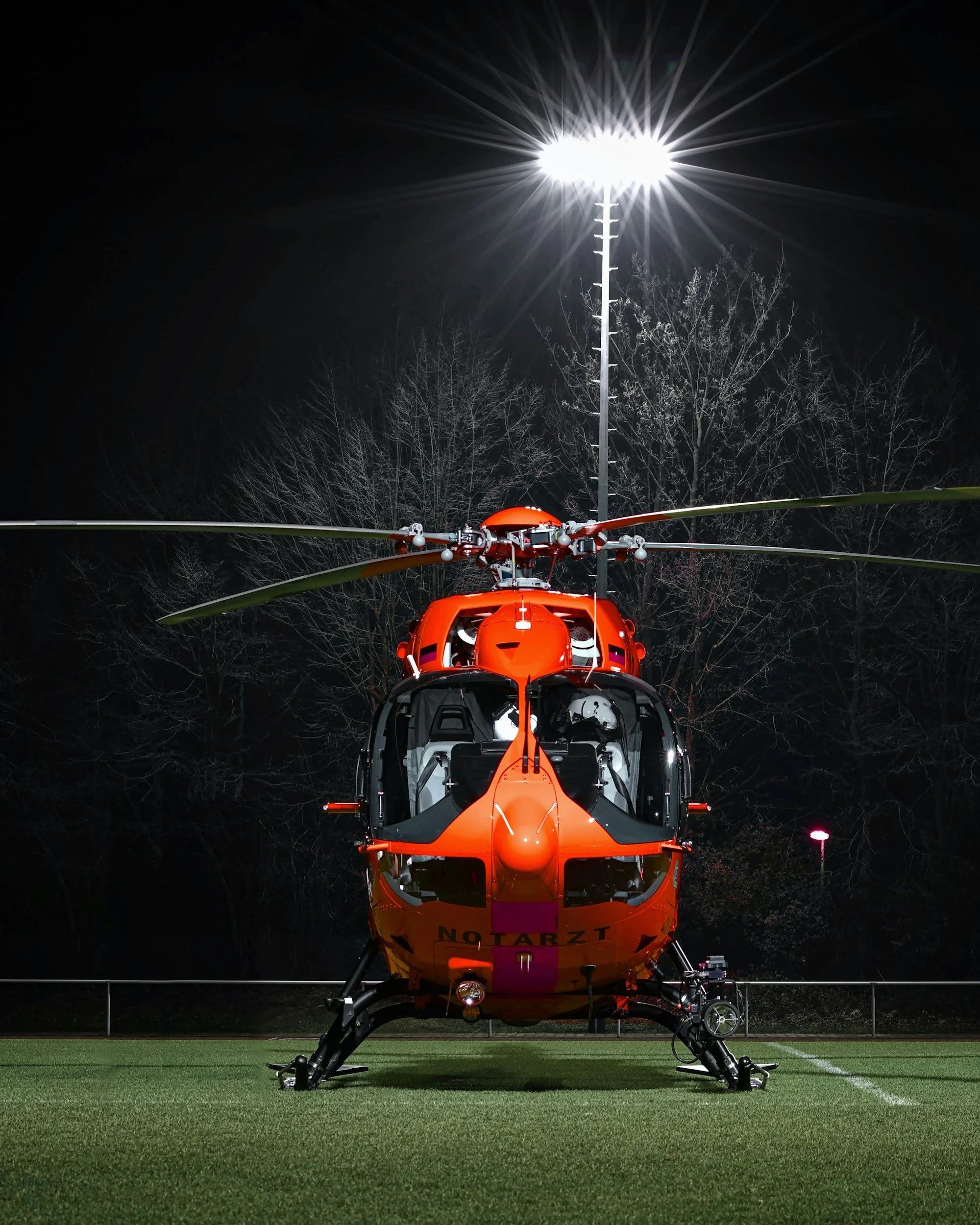 A red emergency helicopter parked on a grassy field at night, illuminated by a tall floodlight, with leafless trees in the background.