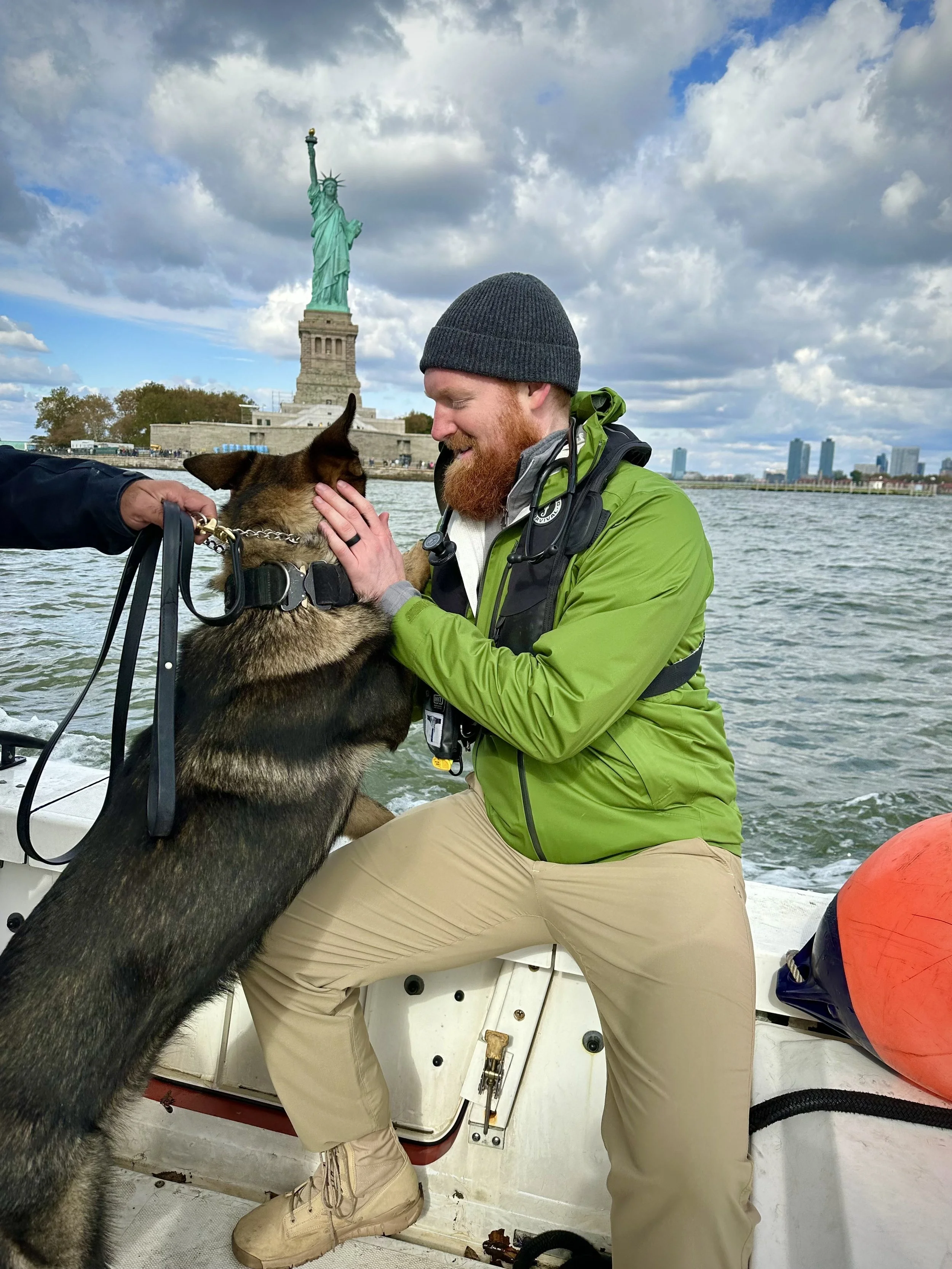 A man in a green jacket and beige pants sitting on a boat, petting a dog with a German Shepherd appearance. The man is smiling and wearing a black beanie. In the background are the Statue of Liberty and a cloudy sky over New York Harbor.