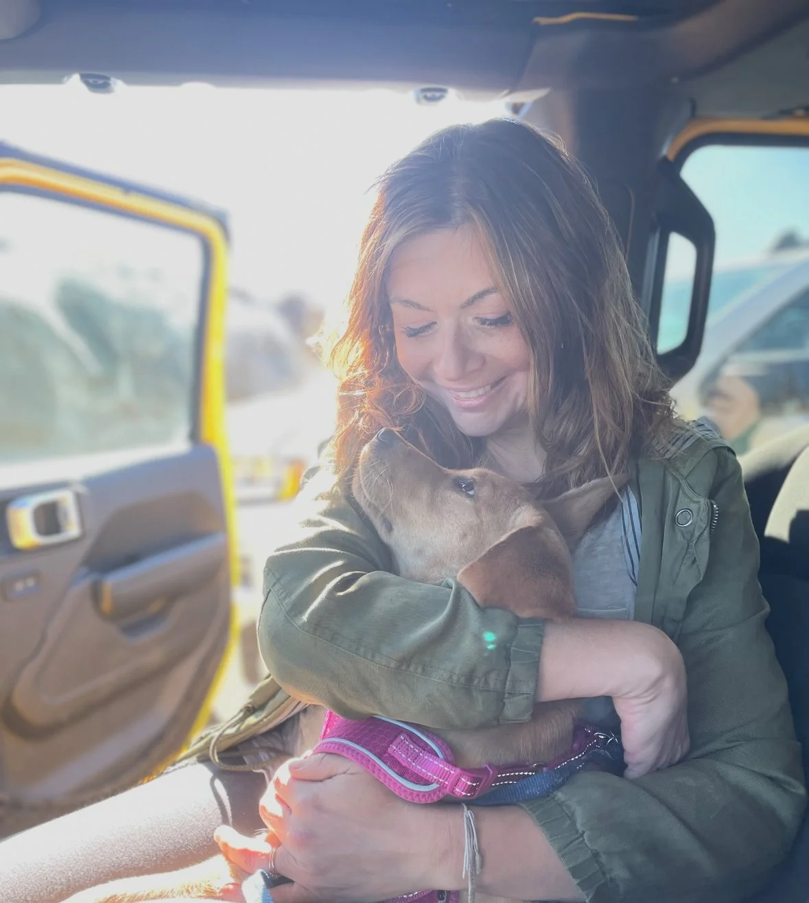 A woman with brown hair smiling while holding a brown puppy in her arms inside a vehicle.