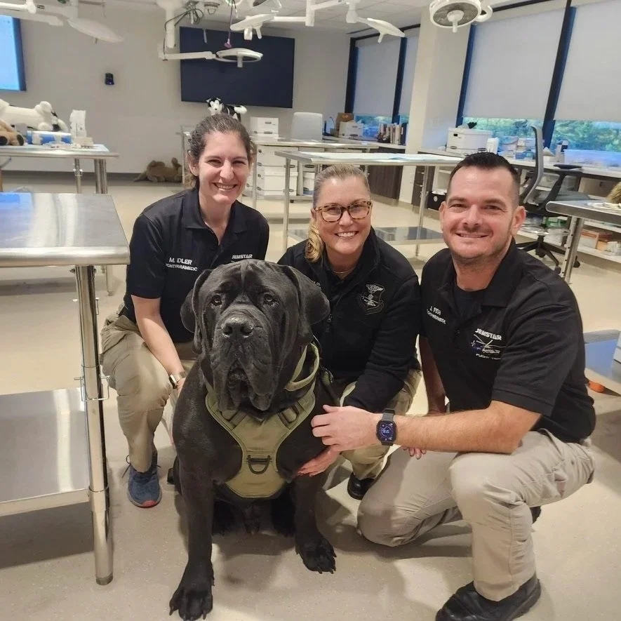 Three people in black shirts with patches and a large black dog wearing a harness, inside a veterinary or medical facility with stainless steel tables and medical equipment.