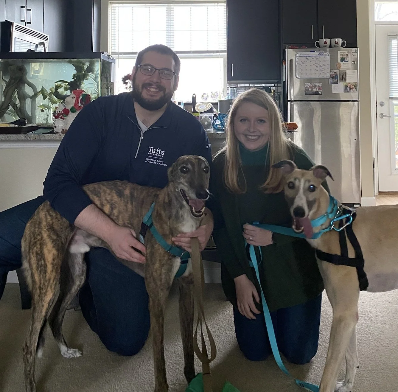 A man and woman smiling with two dogs inside a home, near a kitchen area with cabinets, a refrigerator, and a fish tank in the background.