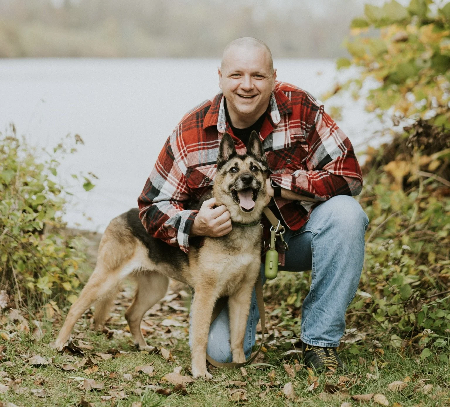 A smiling man crouching next to a happy dog outdoors by a lake, surrounded by fall foliage.