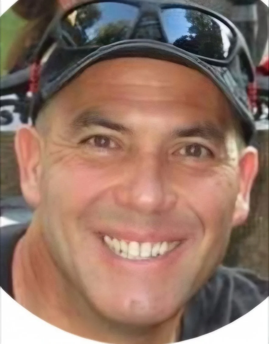 A close-up selfie of a smiling man wearing a cap and sunglasses, with trees and outdoor scenery reflected in the sunglasses.