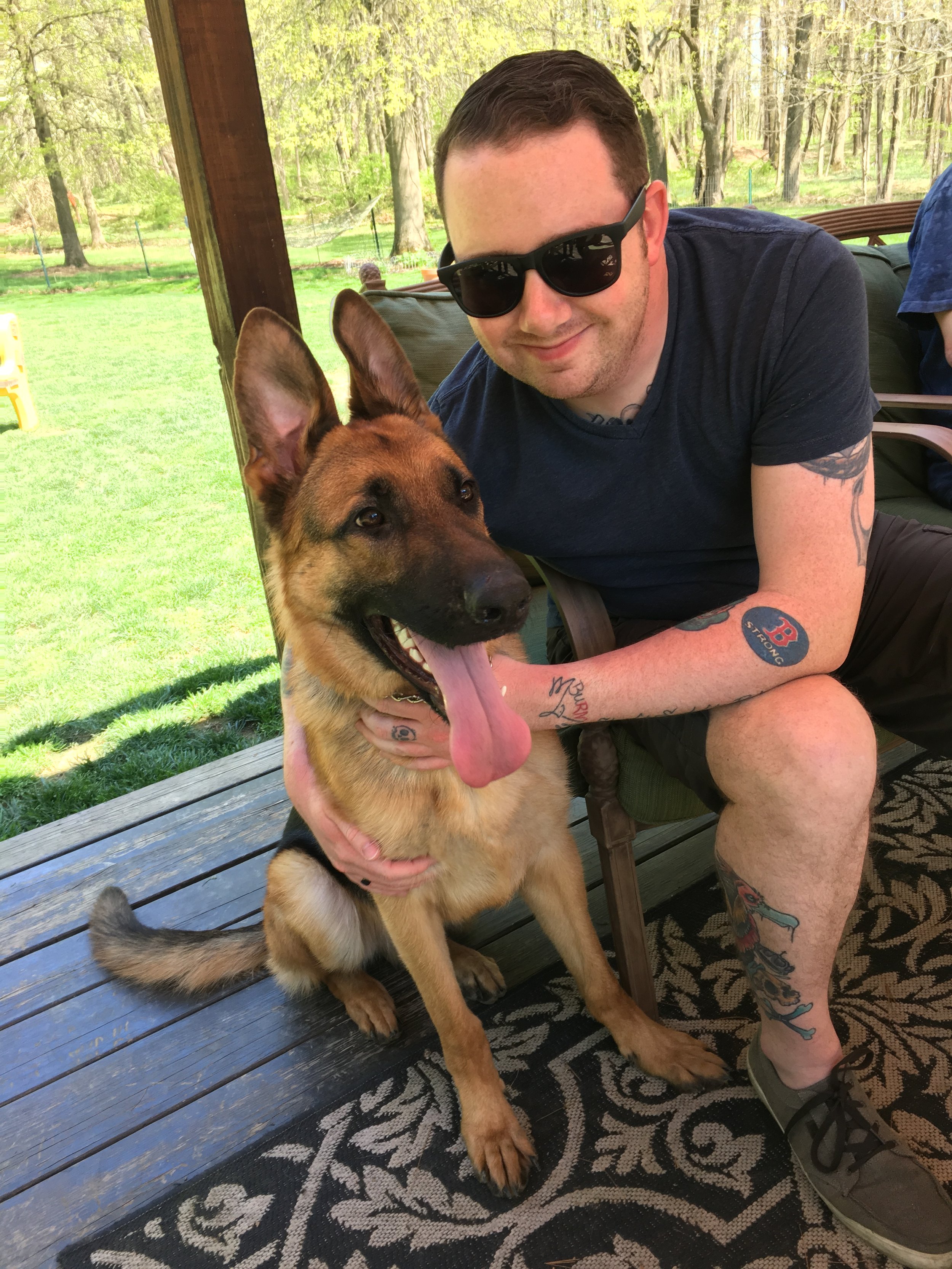 A man with sunglasses and tattoos sitting on a porch next to a large German Shepherd dog with its tongue out in a backyard with trees and grass.