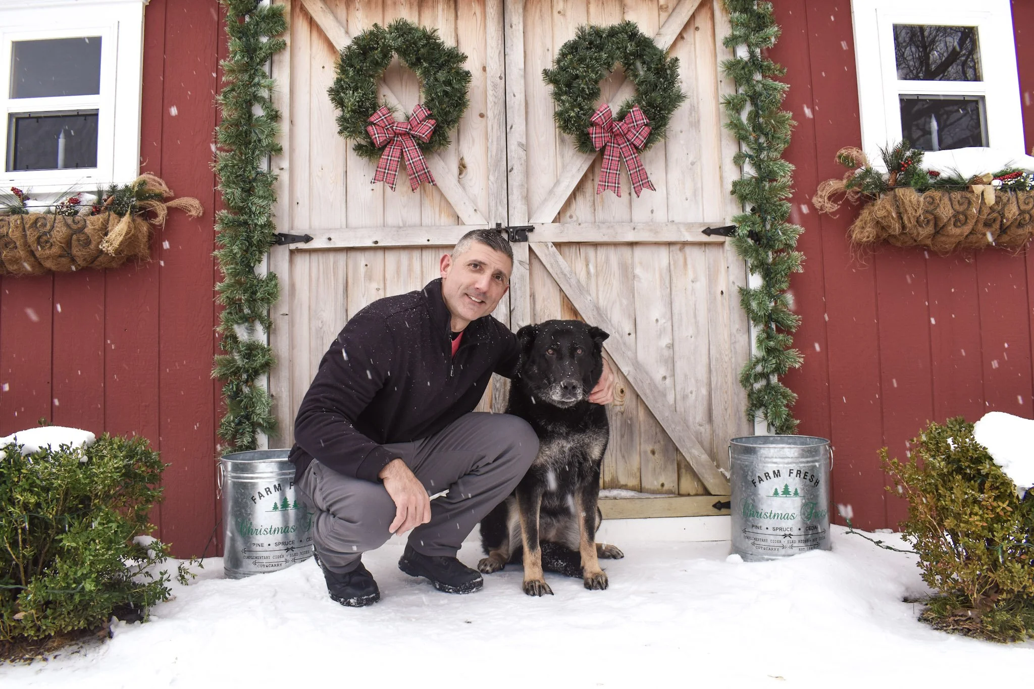 A man and a black and tan dog kneeling in the snow in front of a wooden barn door decorated with Christmas wreaths and green garlands. Snow is falling, and there are bushes and metal buckets with holiday labels on each side.