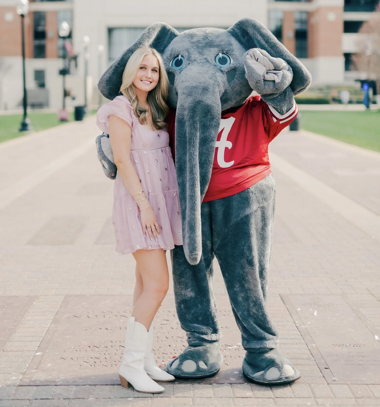 A young woman in a pink dress and white boots standing next to a person dressed in a large gray elephant mascot costume with a red sports jersey, on a paved outdoor area with buildings in the background.