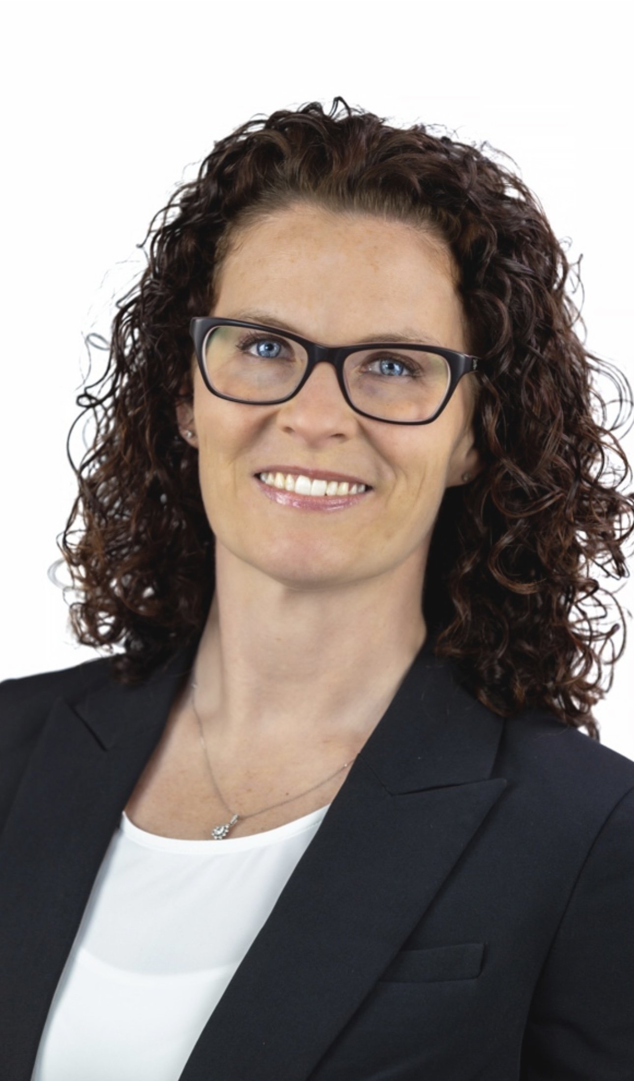 Professional woman with curly brown hair wearing glasses, a black blazer, and a white blouse, smiling against a plain white background.