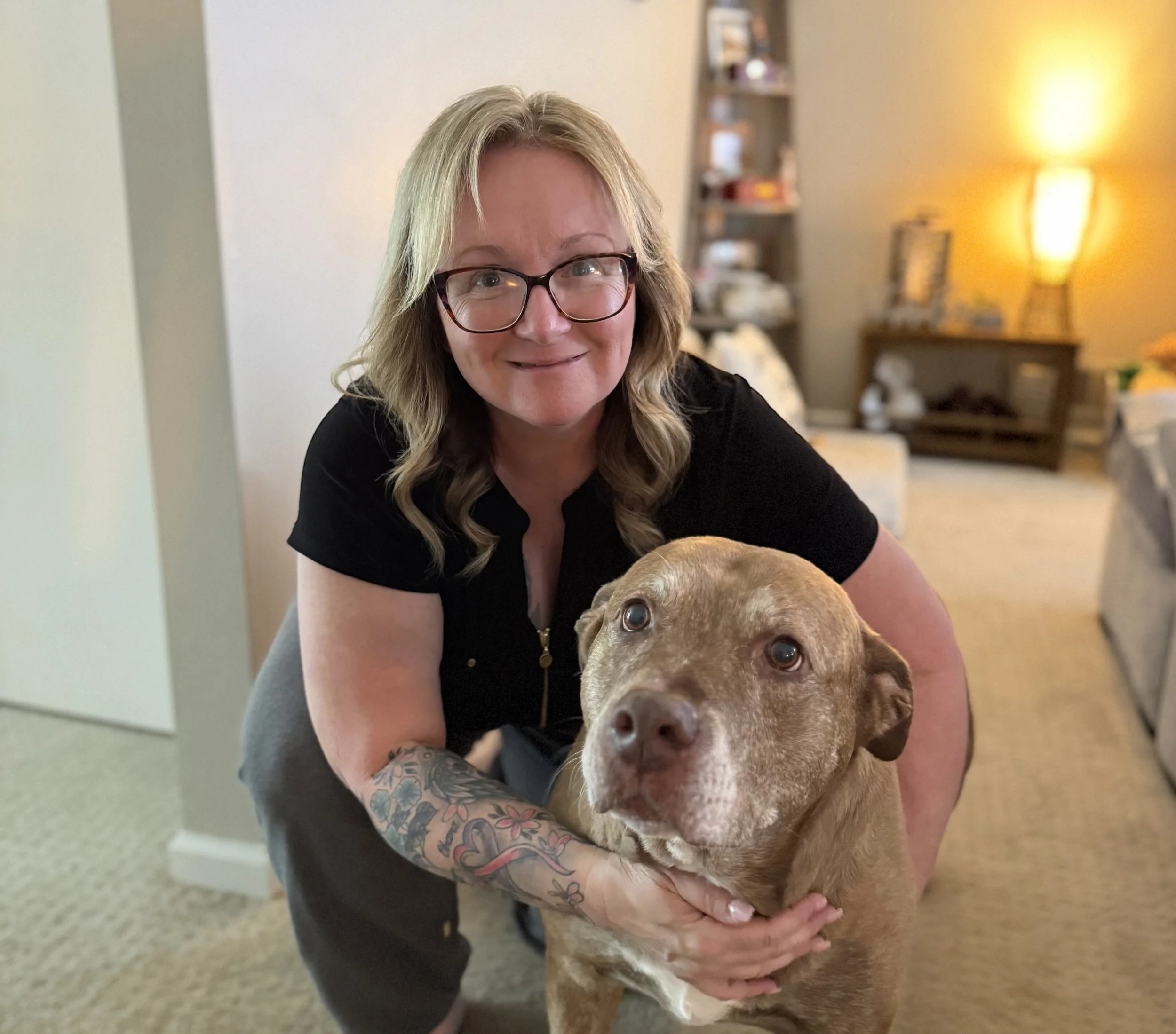 A woman with glasses and tattooed arm smiling while holding a brown dog inside a living room with warm lighting and furniture.