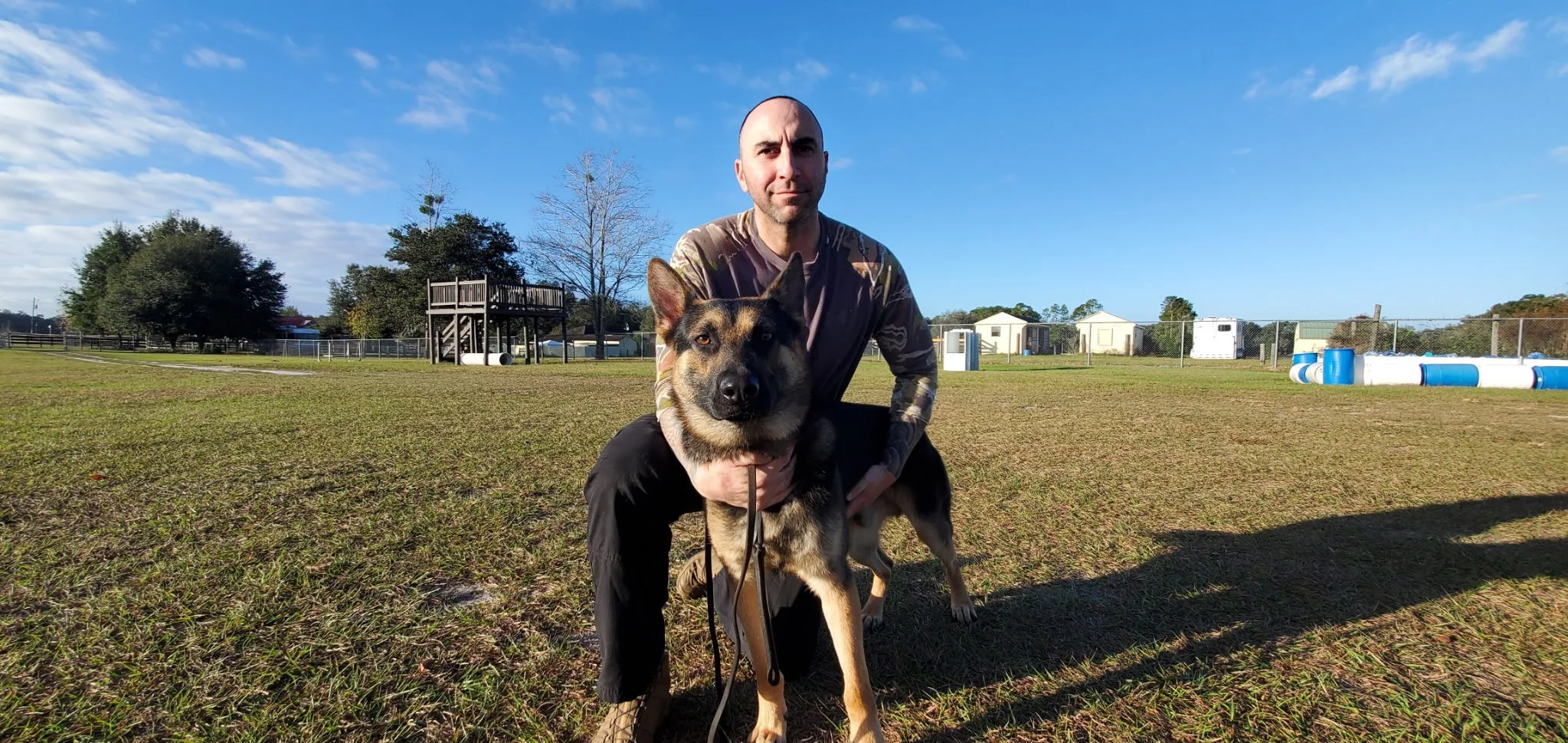 A man in camouflage shirt kneeling next to a large black and brown German Shepherd dog in an open grassy field with blue sky, trees, a wooden structure, and some small buildings in the background.