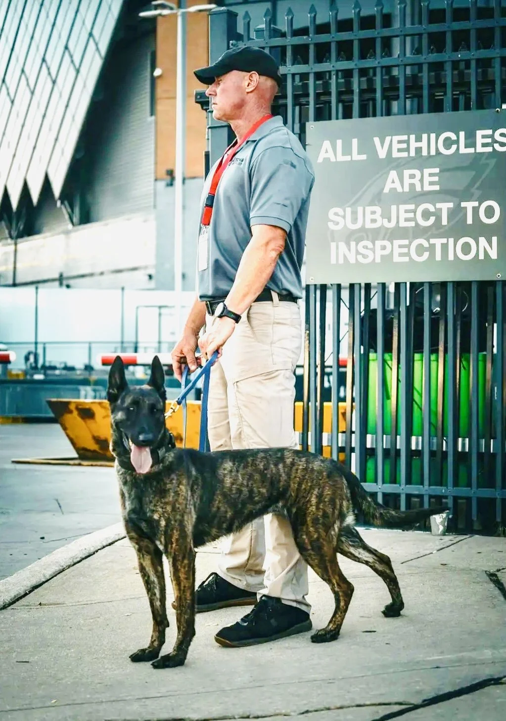 A man in uniform holding a brindle-coated Belgian Malinois dog on a leash at an outdoor vehicle inspection area, with a sign reading 'All vehicles are subject to inspection' in the background.