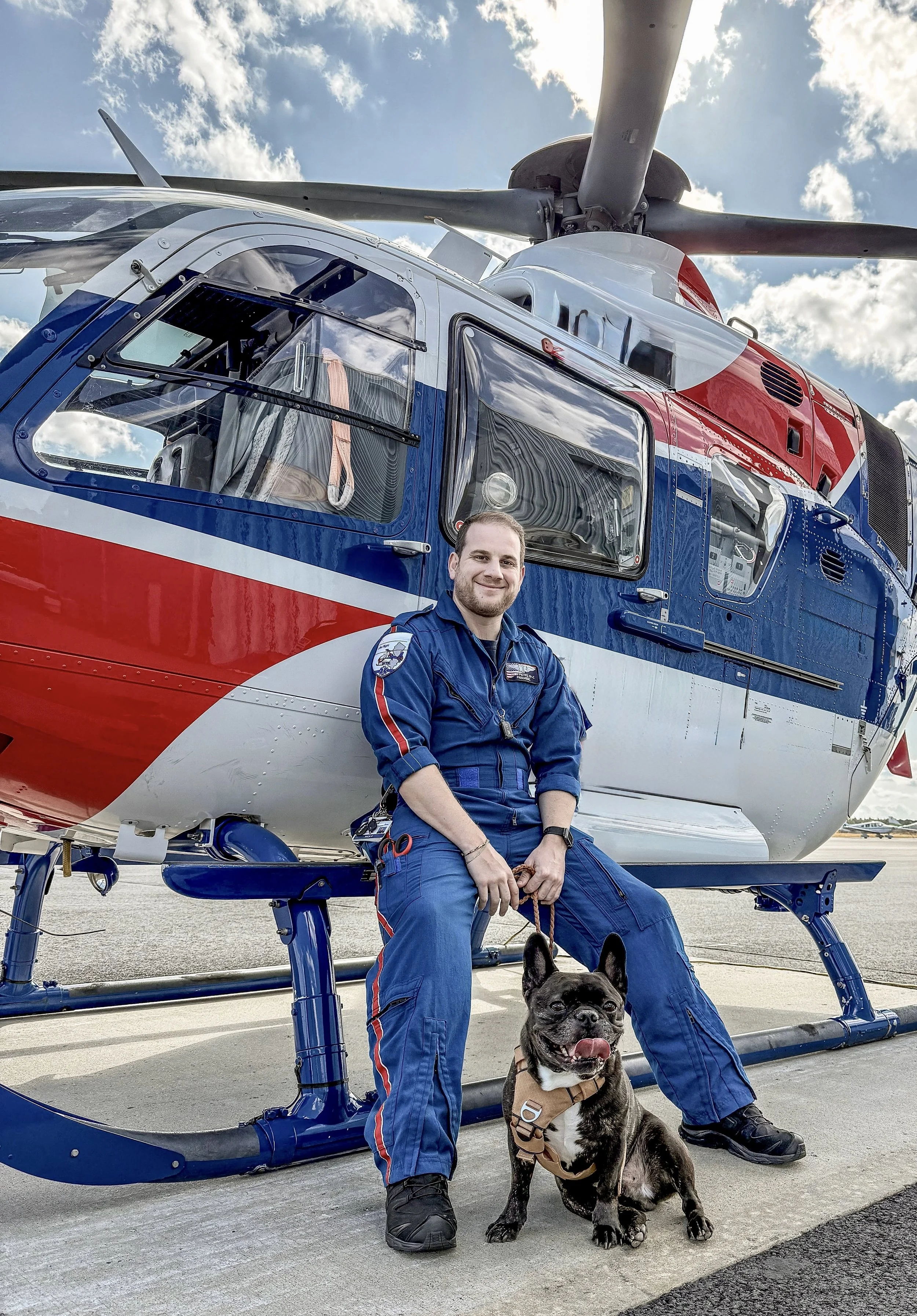 A man in a blue jumpsuit sitting on the landing skids of a helicopter next to his dog, who is also wearing a harness, on an airport tarmac with a partly cloudy sky above.
