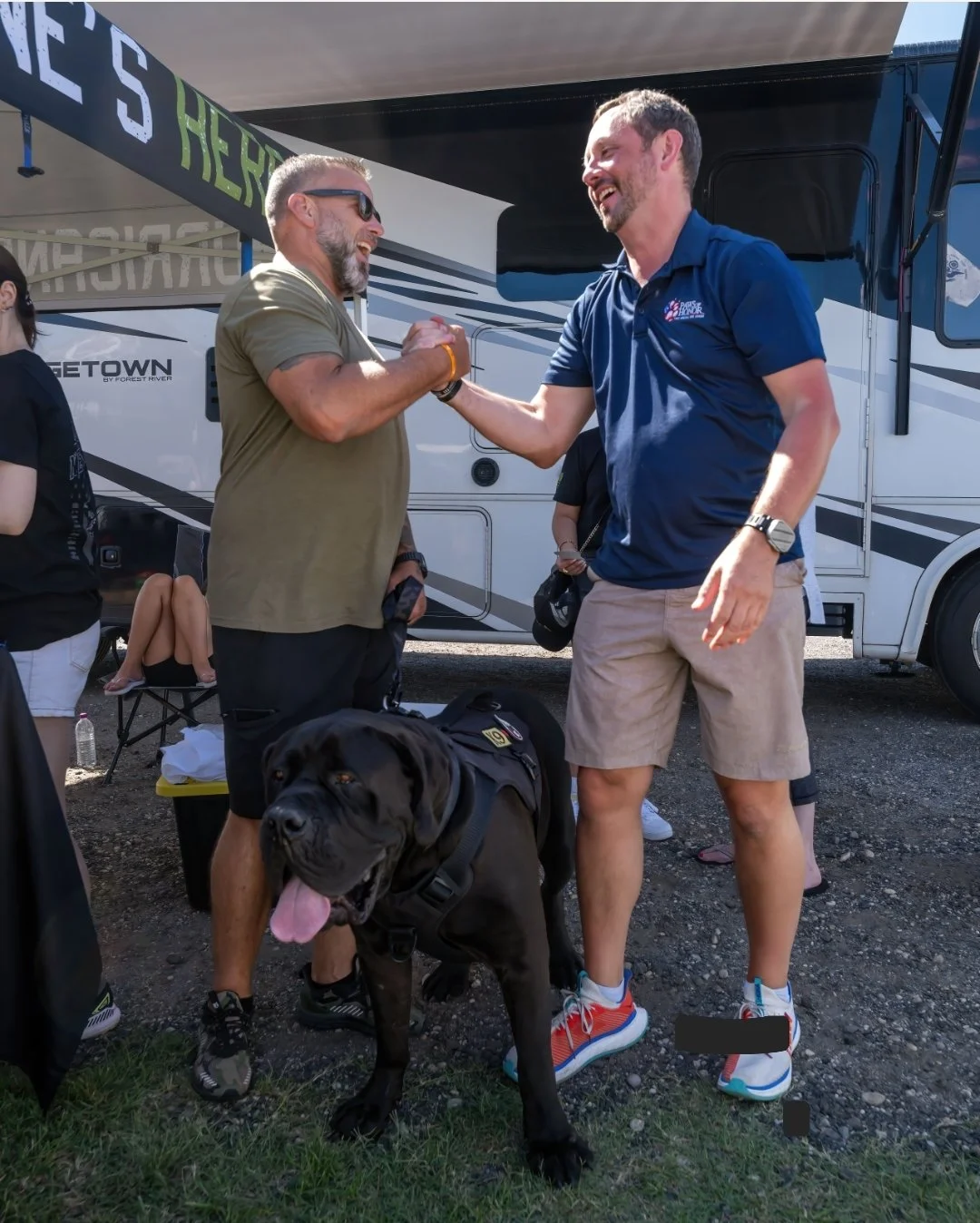 Two men shaking hands and smiling at each other in front of a large RV, with a black dog standing in the foreground. The dog is wearing a harness and sticking out its tongue.