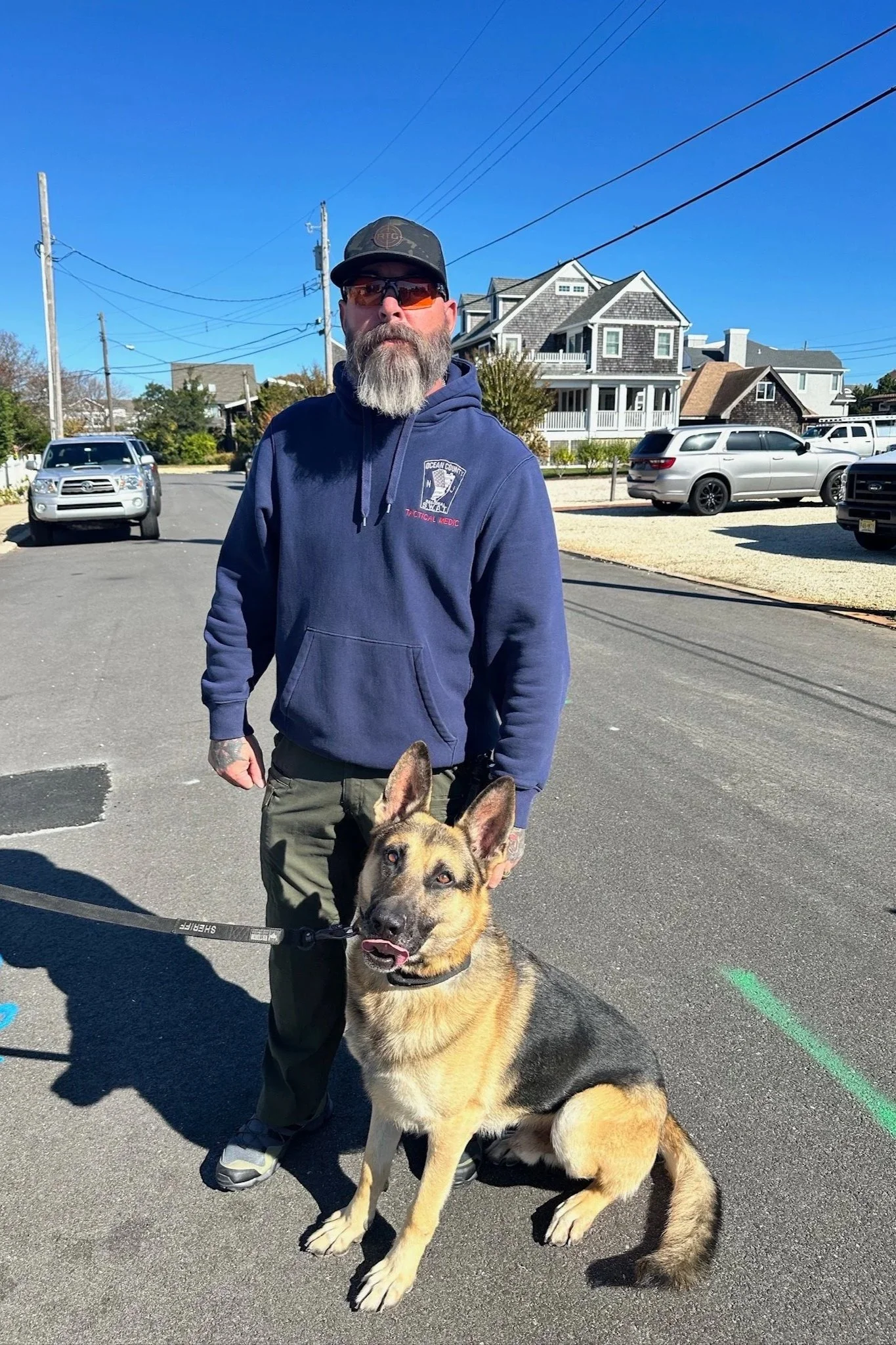 A man with a beard, sunglasses, and a cap standing next to a German Shepherd dog on a leash in a residential neighborhood on a sunny day.