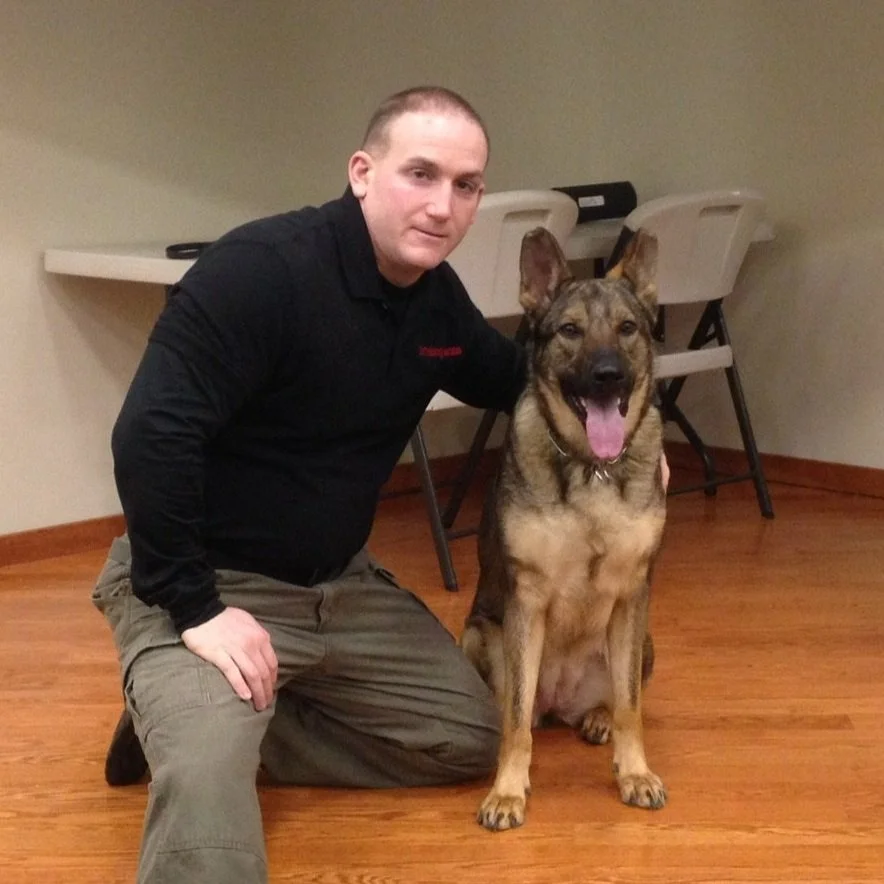 A man kneeling on the floor next to a large, brindle-colored dog in a room with wooden flooring and plain walls, with two white folding chairs in the background.