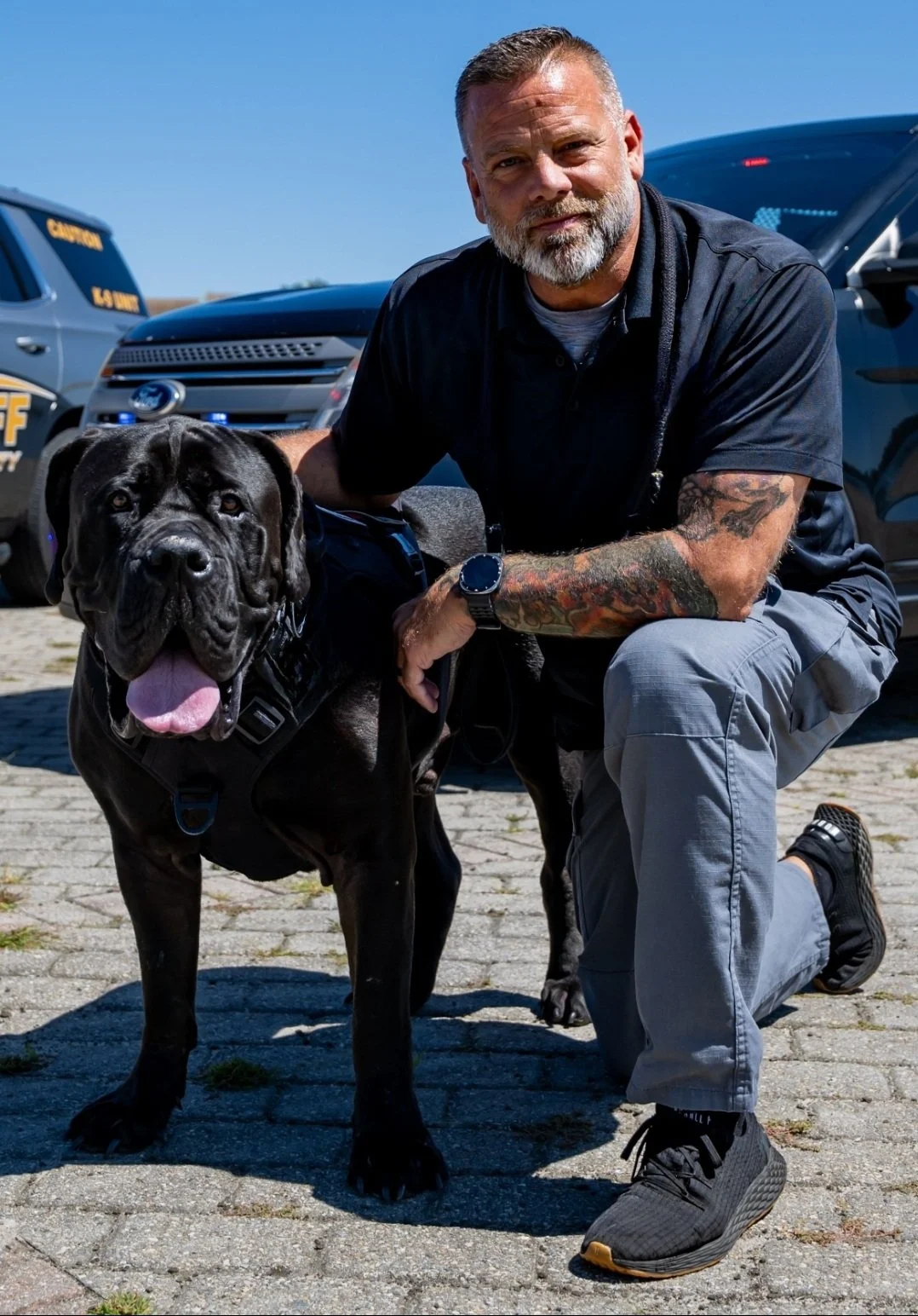A man with tattoos kneeling on one knee next to a large black police dog, both outdoors on a sunny day, with police vehicles in the background.