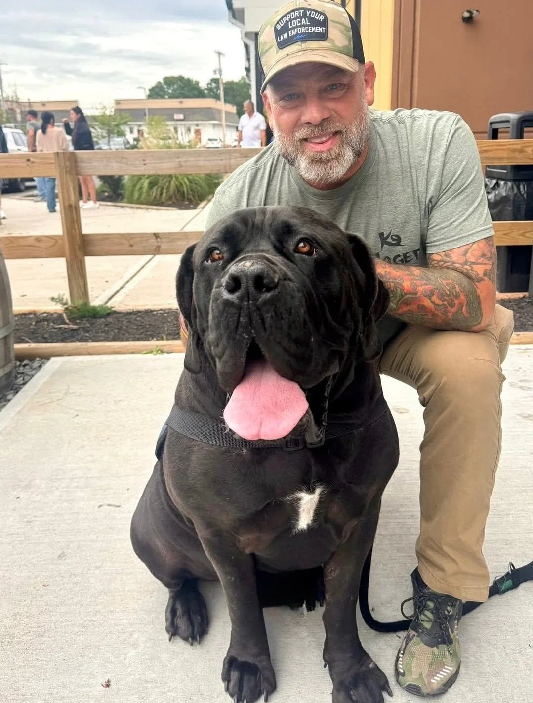 A man with a beard and tattooed arm is squatting next to a large black dog with a broad head, floppy ears, and a pink tongue hanging out, in an outdoor setting with a sidewalk, wooden railing, and people in the background.