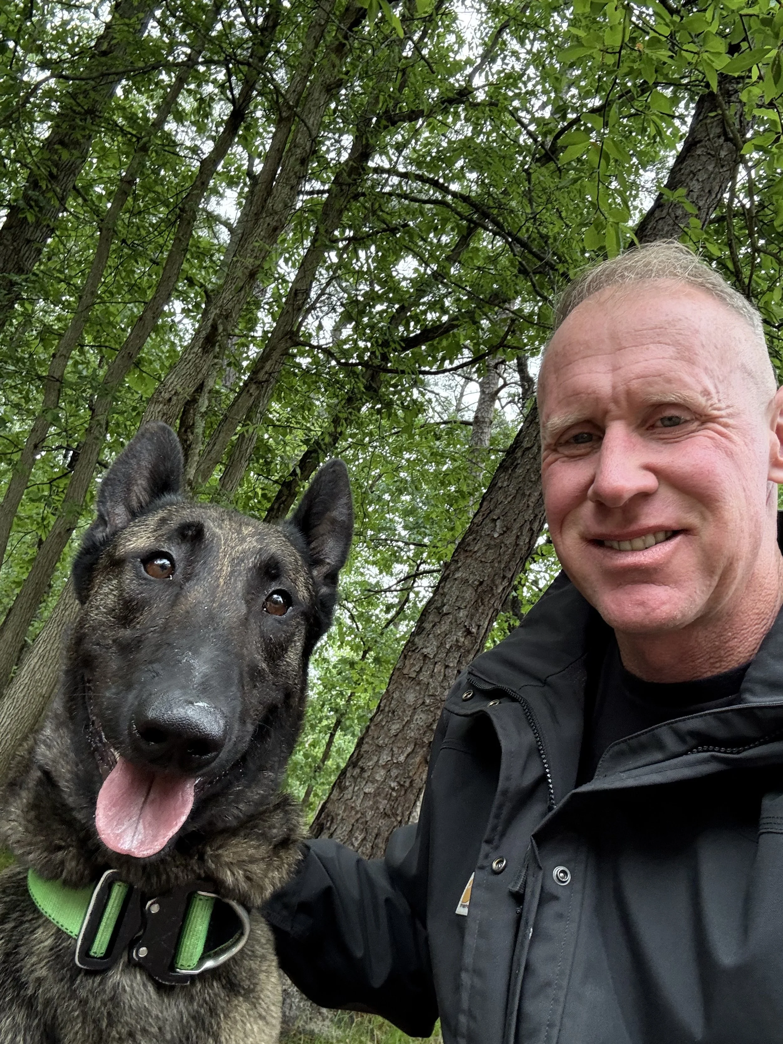A man smiling with a brindle-colored dog in a wooded area with tall trees and green leaves.