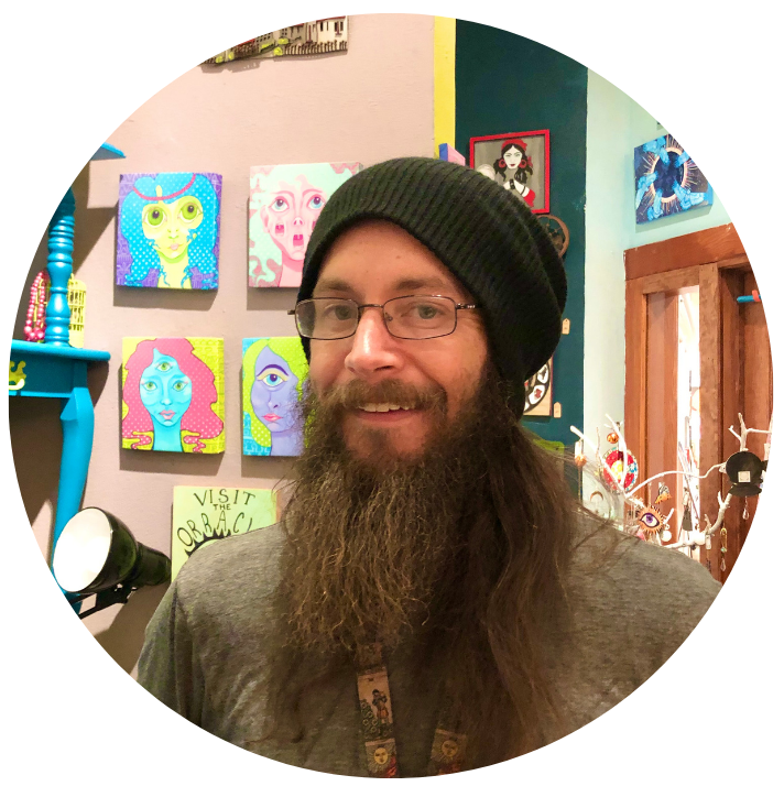 A man with glasses, a long beard, and a black beanie smiling inside a colorful art gallery. The background features vibrant pop art paintings of faces, a sign that says "Visit the Obstacle," and various decorative objects.